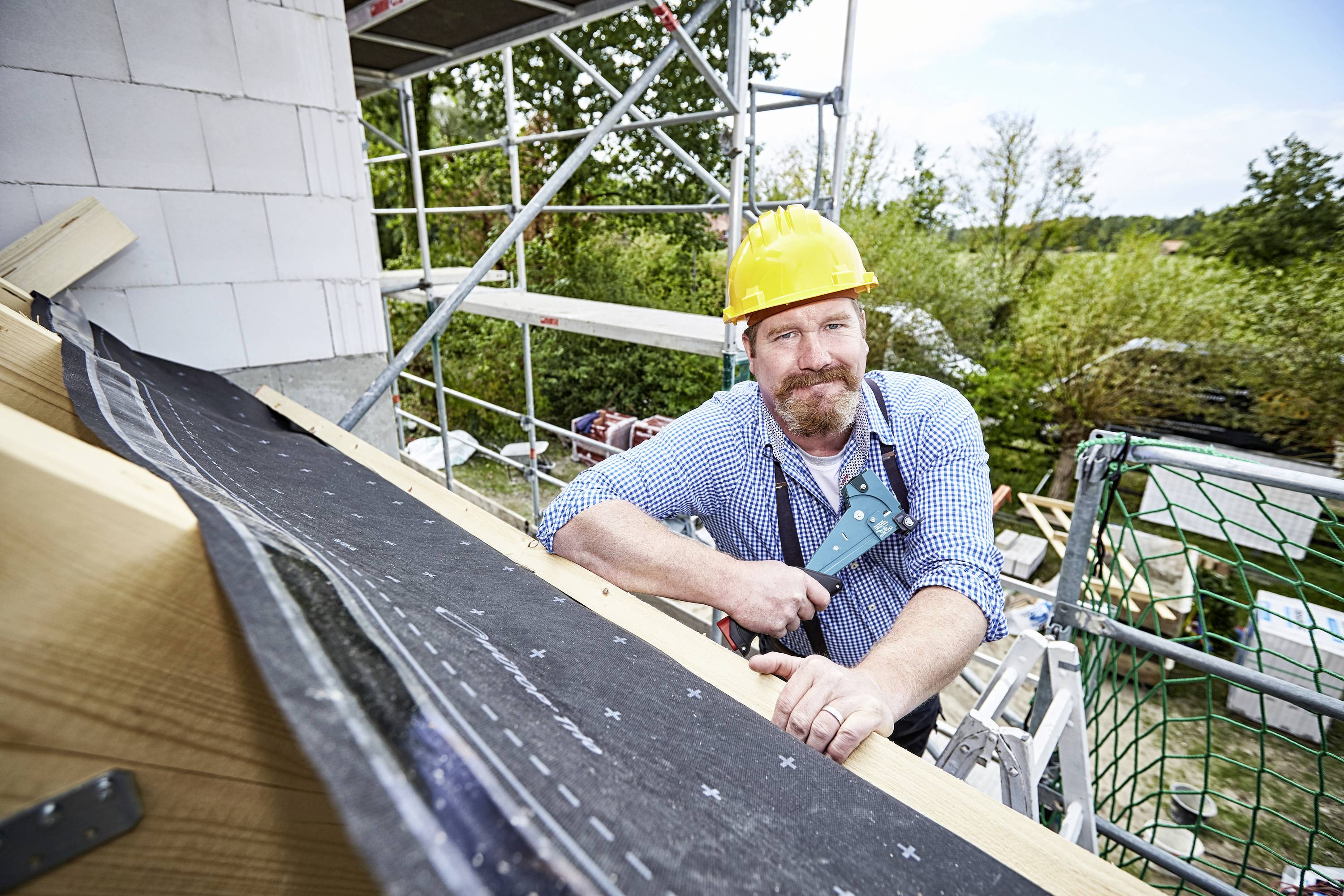Bauarbeiter mit gelbem Helm hält ein Werkzeug und steht auf einem Gerüst, während er an einem Dach eines Hauses arbeitet.