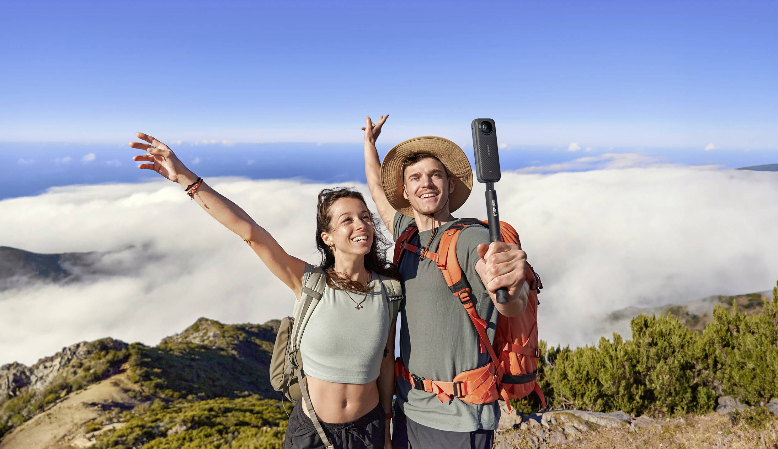 Ein Paar steht auf einem Berggipfel, lächelnd in die Kamera. Sie tragen Rucksäcke und nehmen ein Selfie mit einer malerischen Aussicht auf Wolken und Meer.