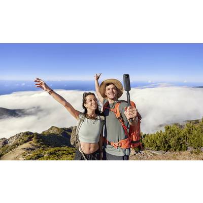 Ein Paar steht auf einem Berggipfel, lächelnd in die Kamera. Sie tragen Rucksäcke und nehmen ein Selfie mit einer malerischen Aussicht auf Wolken und Meer.