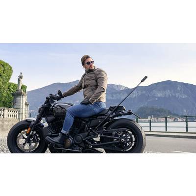 Ein Mann sitzt auf einem Motorrad vor einer Berglandschaft. Er trägt eine Sonnenbrille und eine braune Jacke.