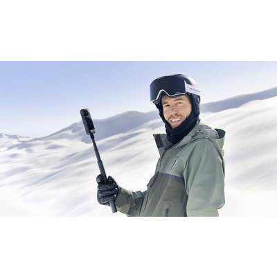 Ein Mensch in Winterkleidung hält einen Selfie-Stick und lächelt in einer schneebedeckten Berglandschaft.