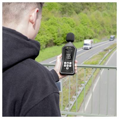 Person mit einem Schallpegelmesser zur Messung von Verkehrslärm neben einer Autobahn, mit vorbeifahrenden Lastkraftwagen im Hintergrund.