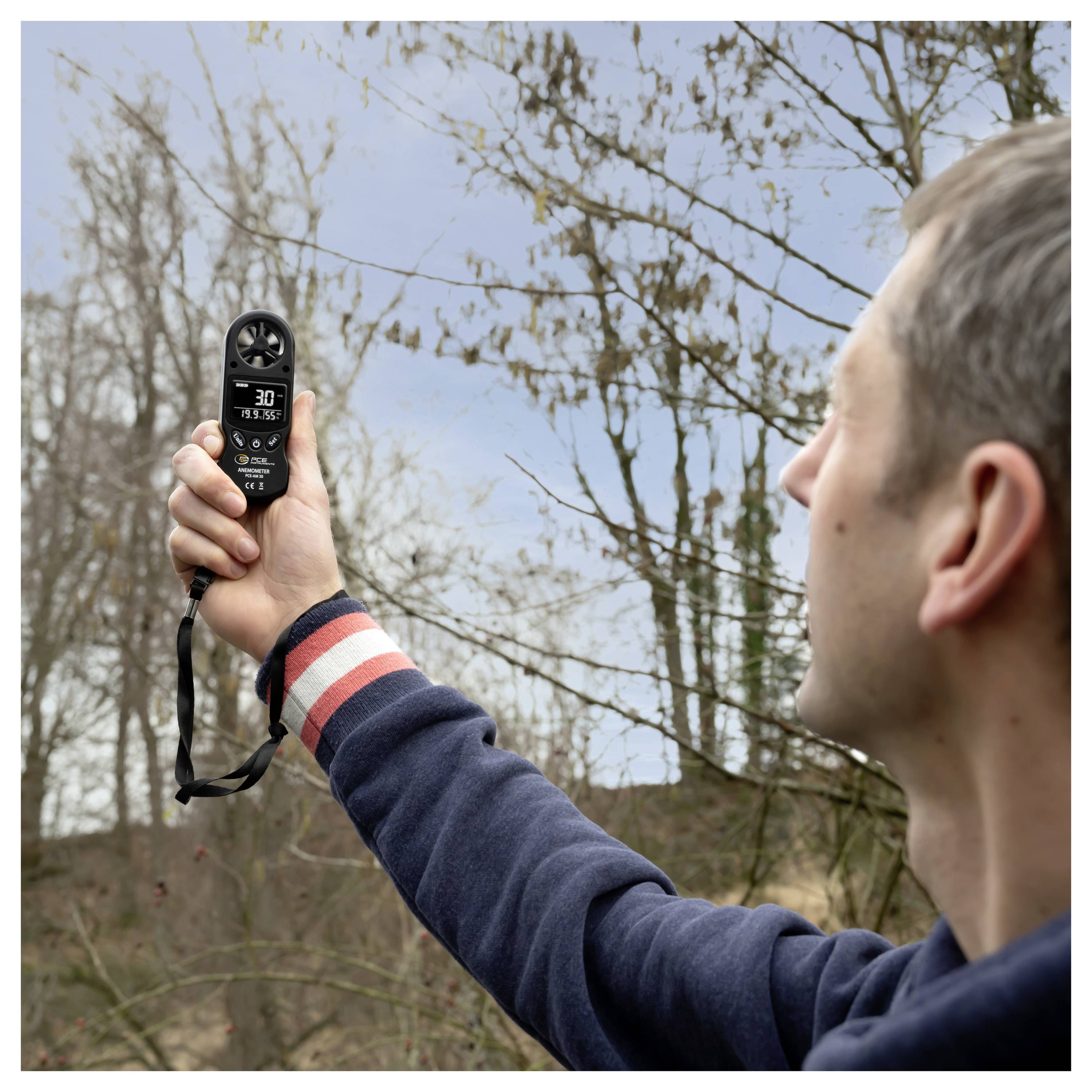 Mann im Freien, der die Windgeschwindigkeit mit einem tragbaren Anemometer misst, hält es zu kahlen Bäumen unter einem bewölkten Himmel hoch.