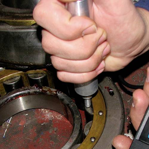 Eine Hand hält ein Werkzeug und bearbeitet ein Metalllager. Im Hintergrund sind Zahnräder sichtbar, die auf eine Werkstattumgebung hindeuten.