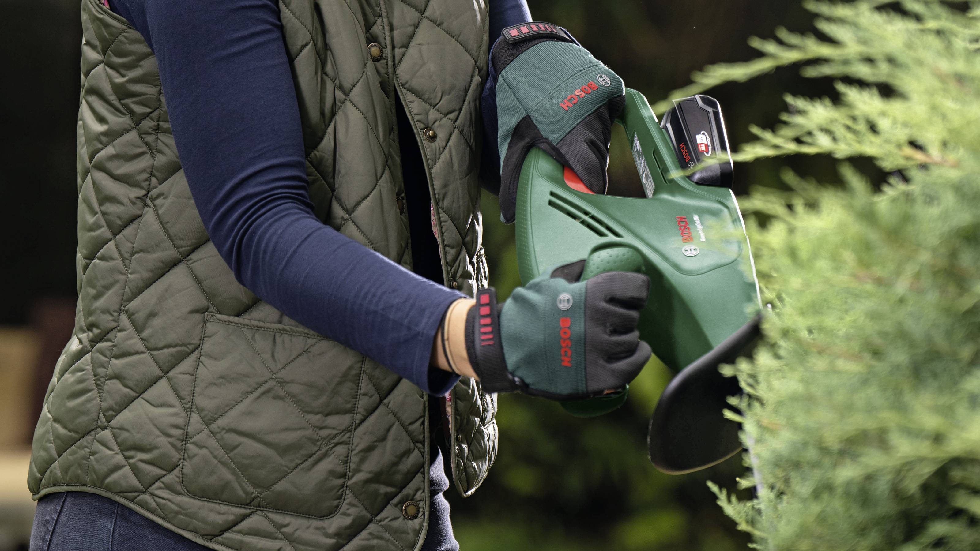 Eine Person schneidet mit einem elektrischen Heckenschneider eine Hecke. Sie trägt eine grüne Weste und Arbeitshandschuhe.