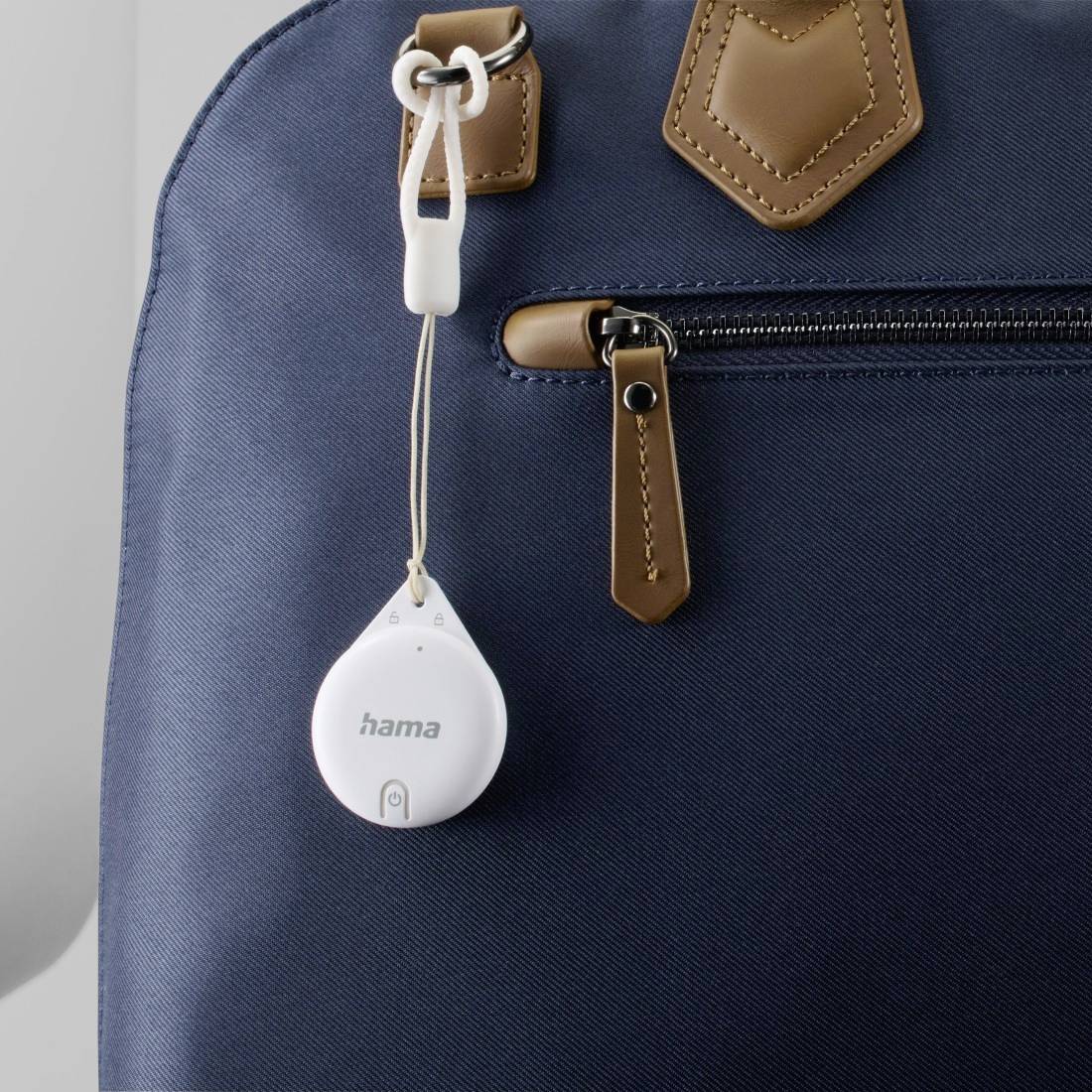 Ein runder, weißer Tracker hängt an einer blauen Tasche aus Stoff mit braunen Lederdetails.