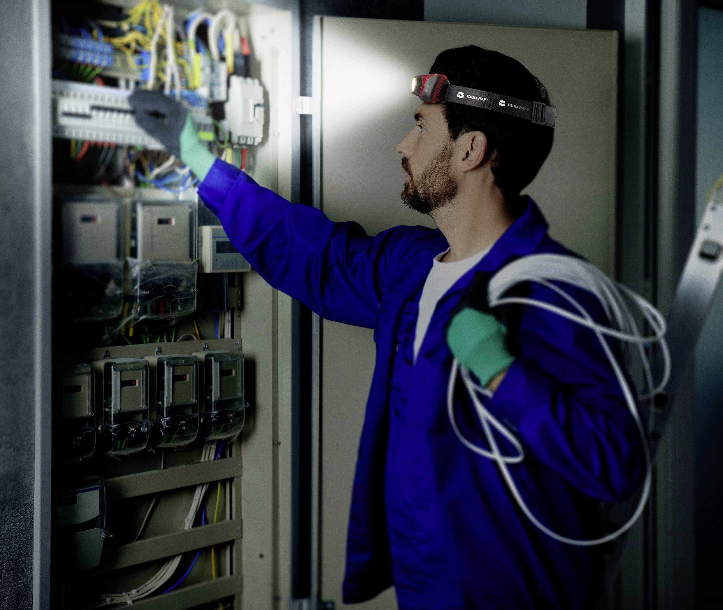 Ein Elektriker in blauer Arbeitskleidung überprüft einen Schaltschrank und hält dabei ein Kabelbündel.