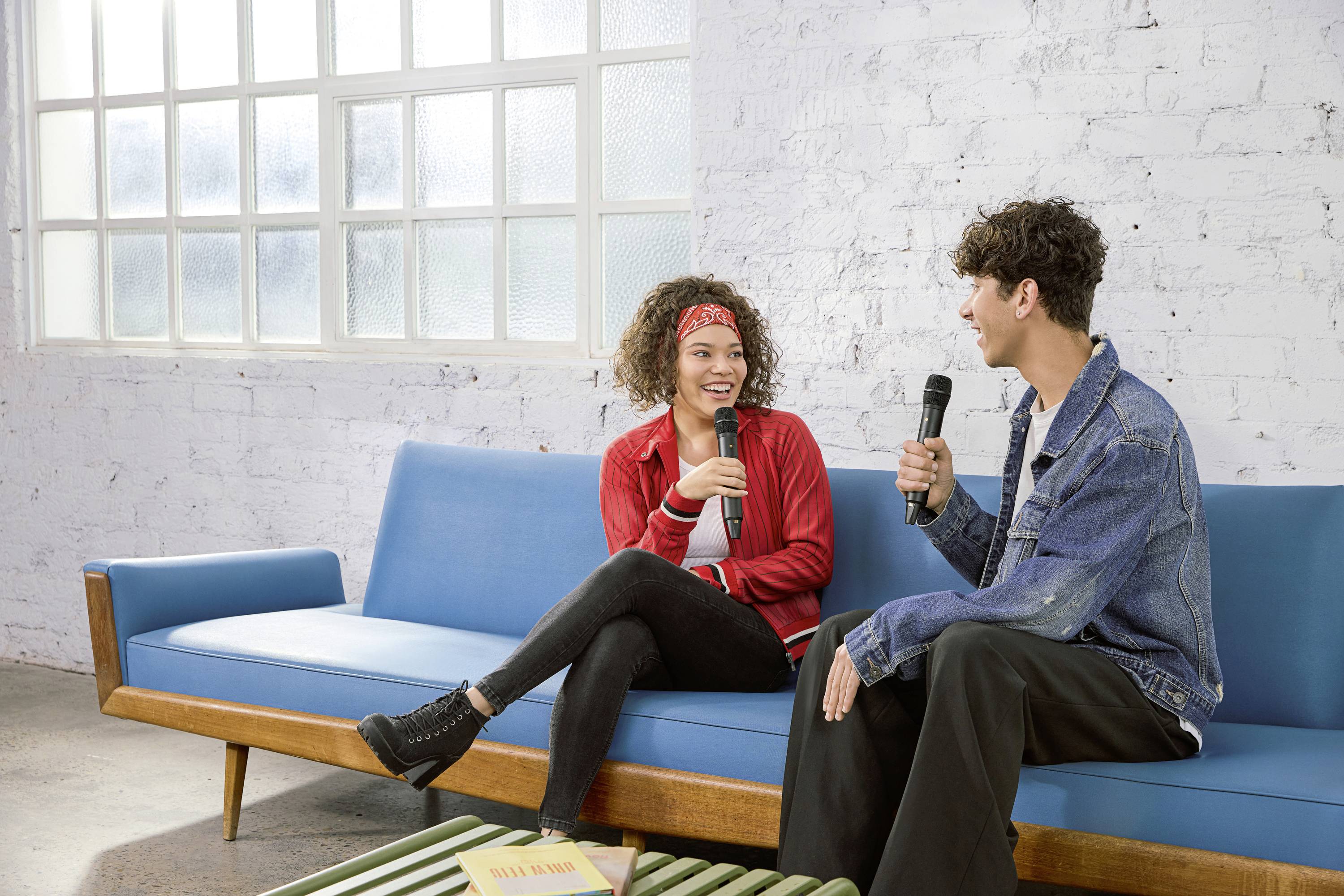 Zwei Personen sitzen auf einem blauen Sofa und sprechen fröhlich in Mikrofone. Im Hintergrund ist eine helle, gemauerte Wand.