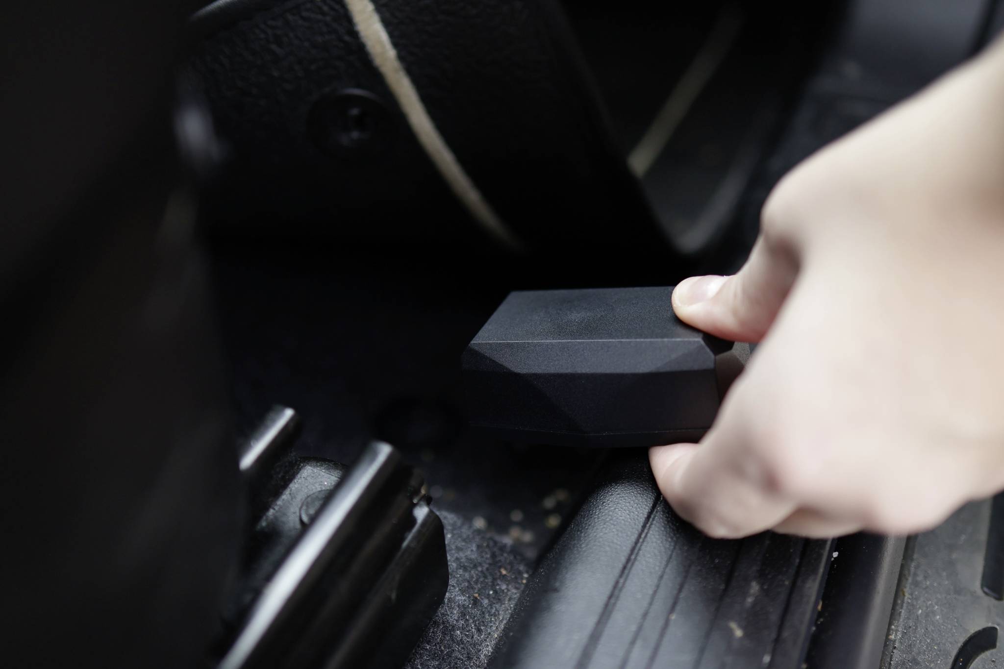 Eine Person hält ein kleines, schwarzes Gerät in der Hand und platziert es in den Fußraum eines Fahrzeugs, nahe dem Sitzgestell.
