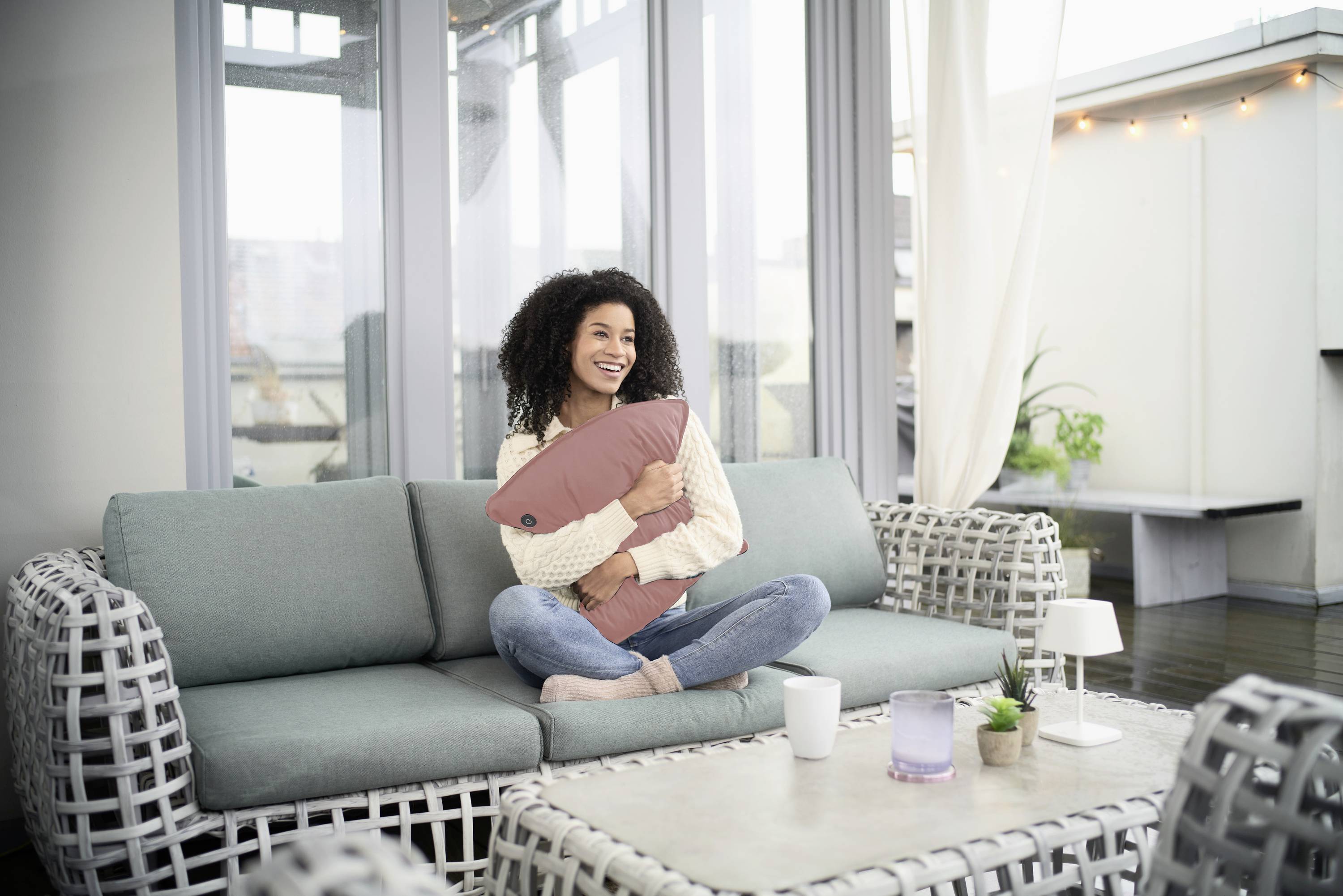 Frau sitzt lächelnd auf einem Sofa, hält ein Kissen in den Armen. Sie befindet sich in einem hell eingerichteten Raum mit Pflanzen im Hintergrund.