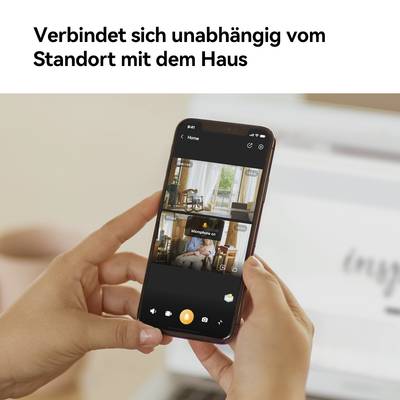 Eine Hand hält ein Smartphone mit einer App zur Überwachung von Innenräumen. Text oben: 'Verbindet sich unabhängig vom Standort mit dem Haus'.