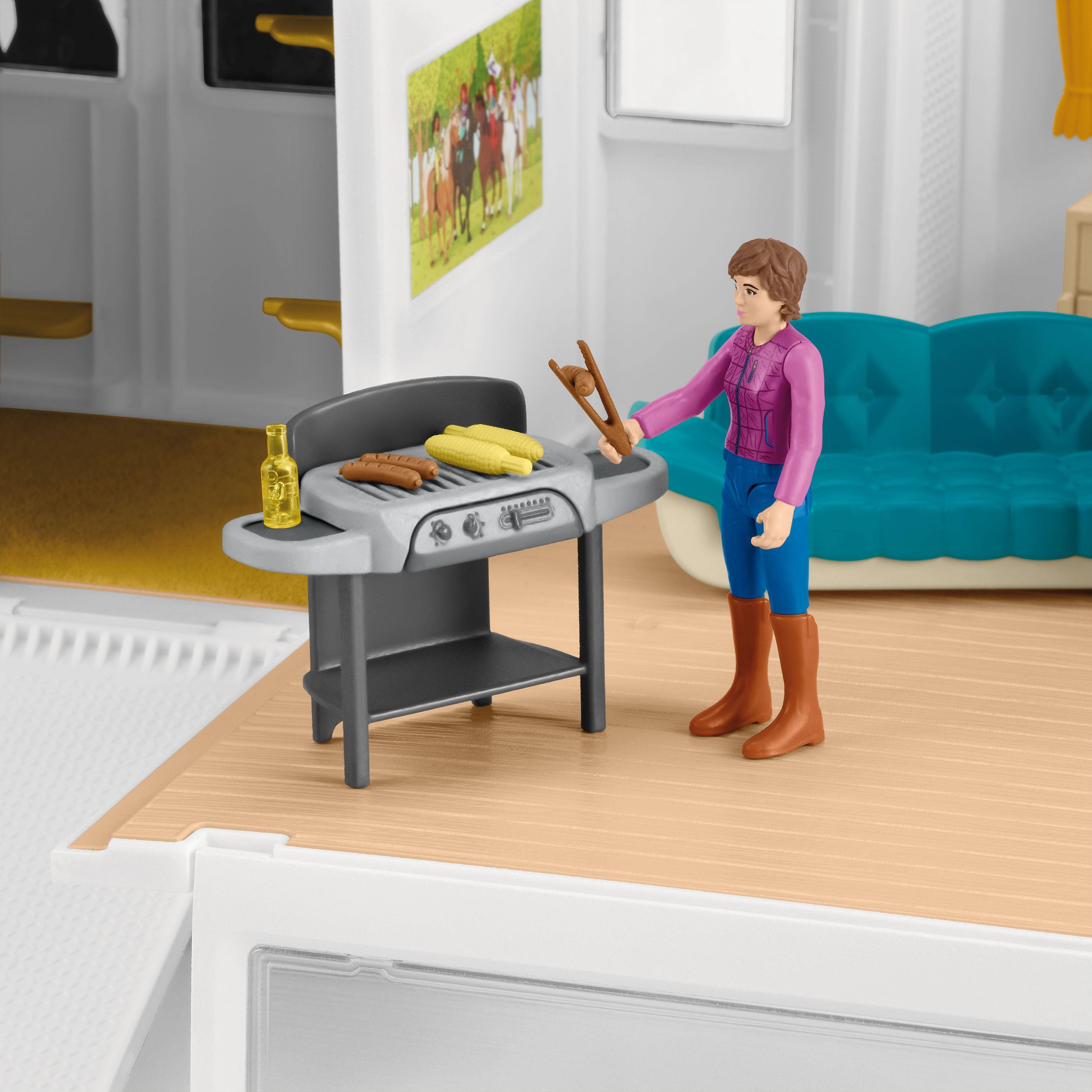 Eine Figur steht in einem Puppenhaus neben einem Tisch mit einem Grill und Essen. Im Hintergrund sind ein Sofa und ein Bild an der Wand.