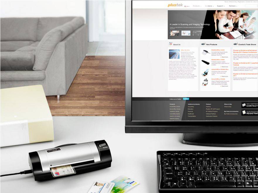 Büroarbeitsplatz mit Scanner und Computermonitor. Der Monitor zeigt eine Webseite mit Informationen zu Scan- und Imaging-Technologie. Im Hintergrund ist ein Sofa zu sehen.