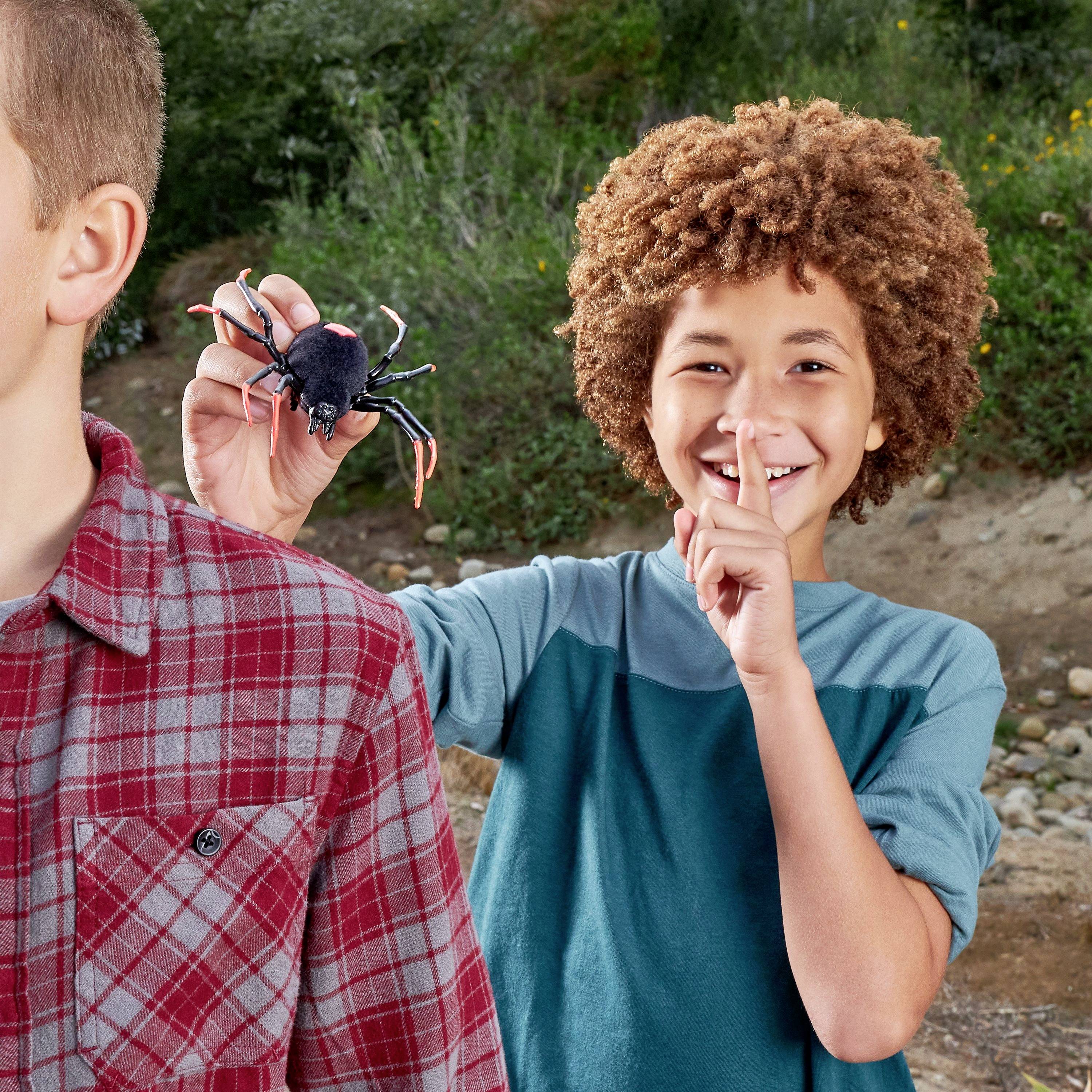 Ein Kind zeigt einem anderen Kind eine Spielzeugspinne als Scherz, während das andere überrascht schaut. Hintergrund: Natur.