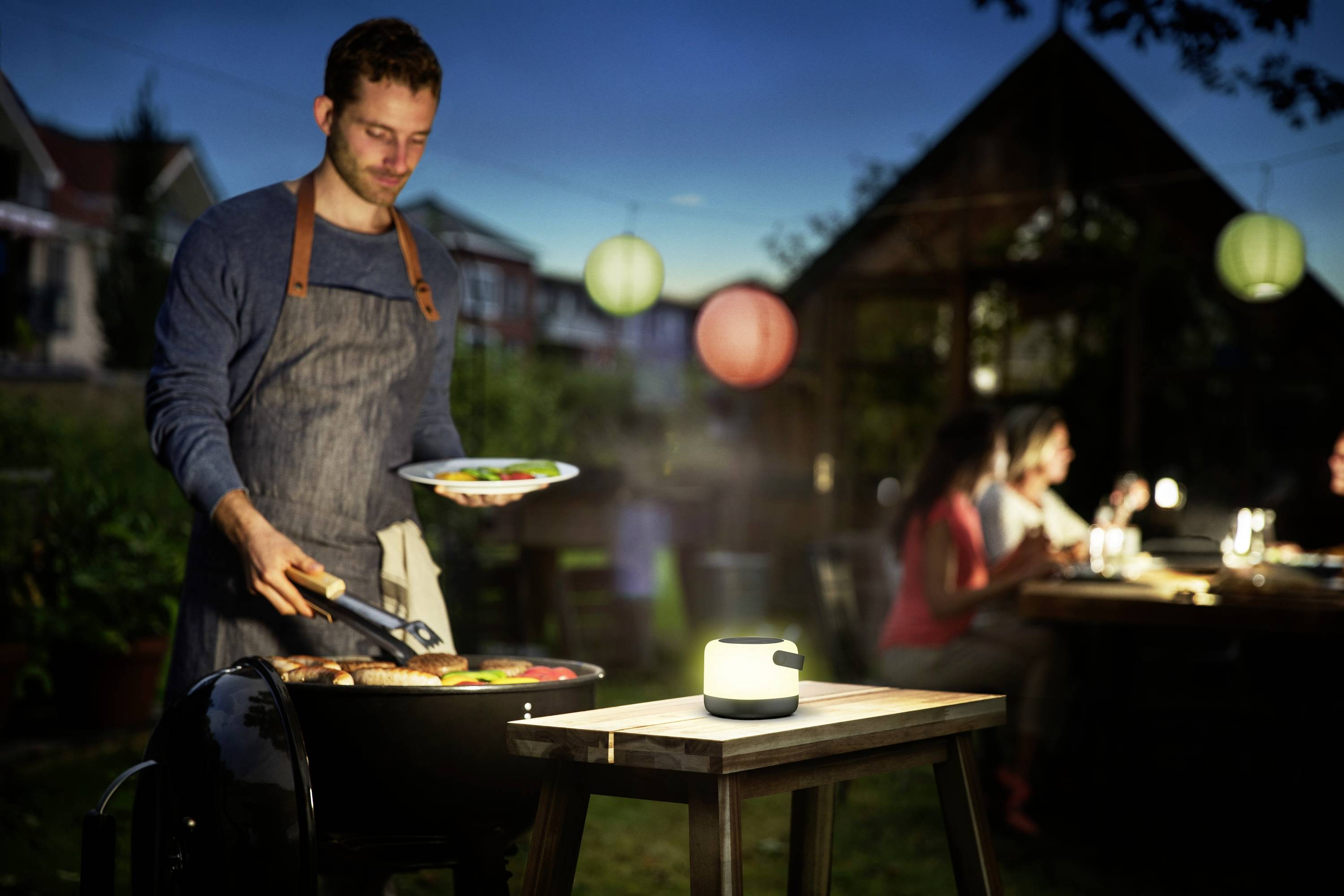 Ein Mann grillt Fleisch und Gemüse im Garten am Abend, während im Hintergrund Menschen an einem Tisch sitzen und essen.