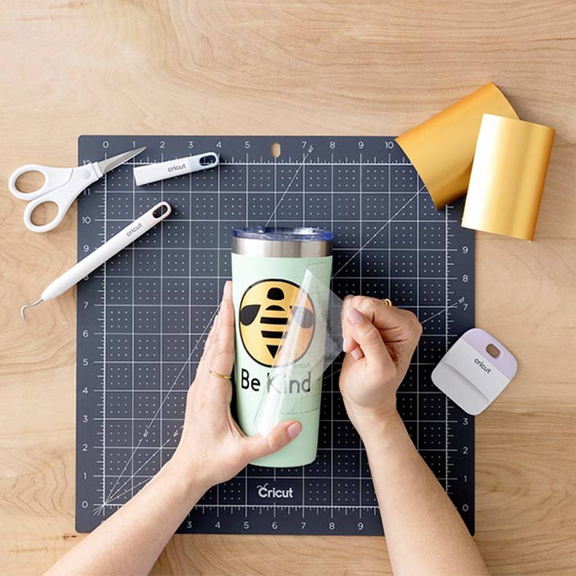 Auf einer Schneidematte wird ein grüner Becher mit einem 'Be Kind'-Logo aus Vinylfolie verziert. Werkzeuge und Folien sind daneben platziert.