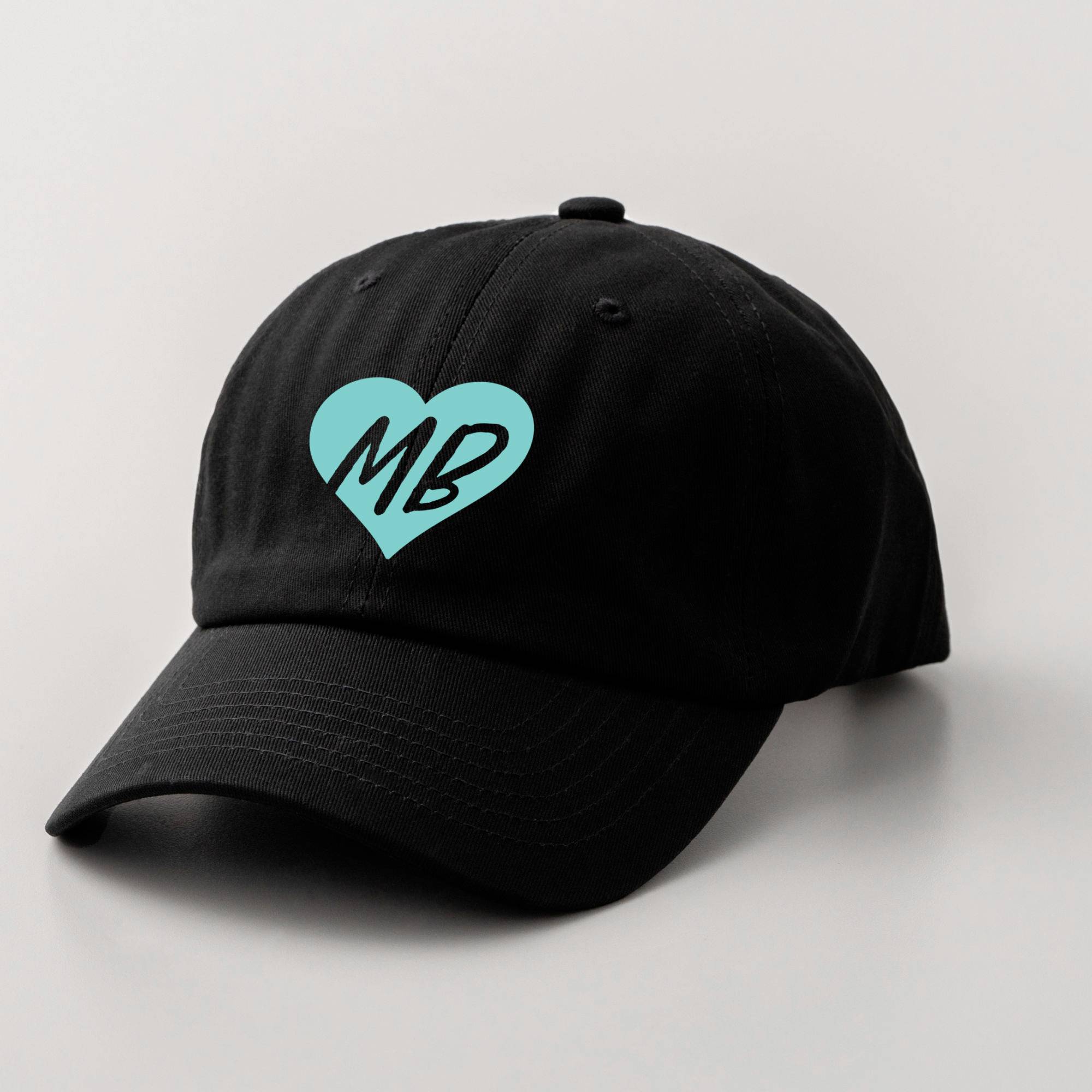 Schwarze Baseballkappe mit einem türkisfarbenen Herz und den Initialen 'MB' darauf.