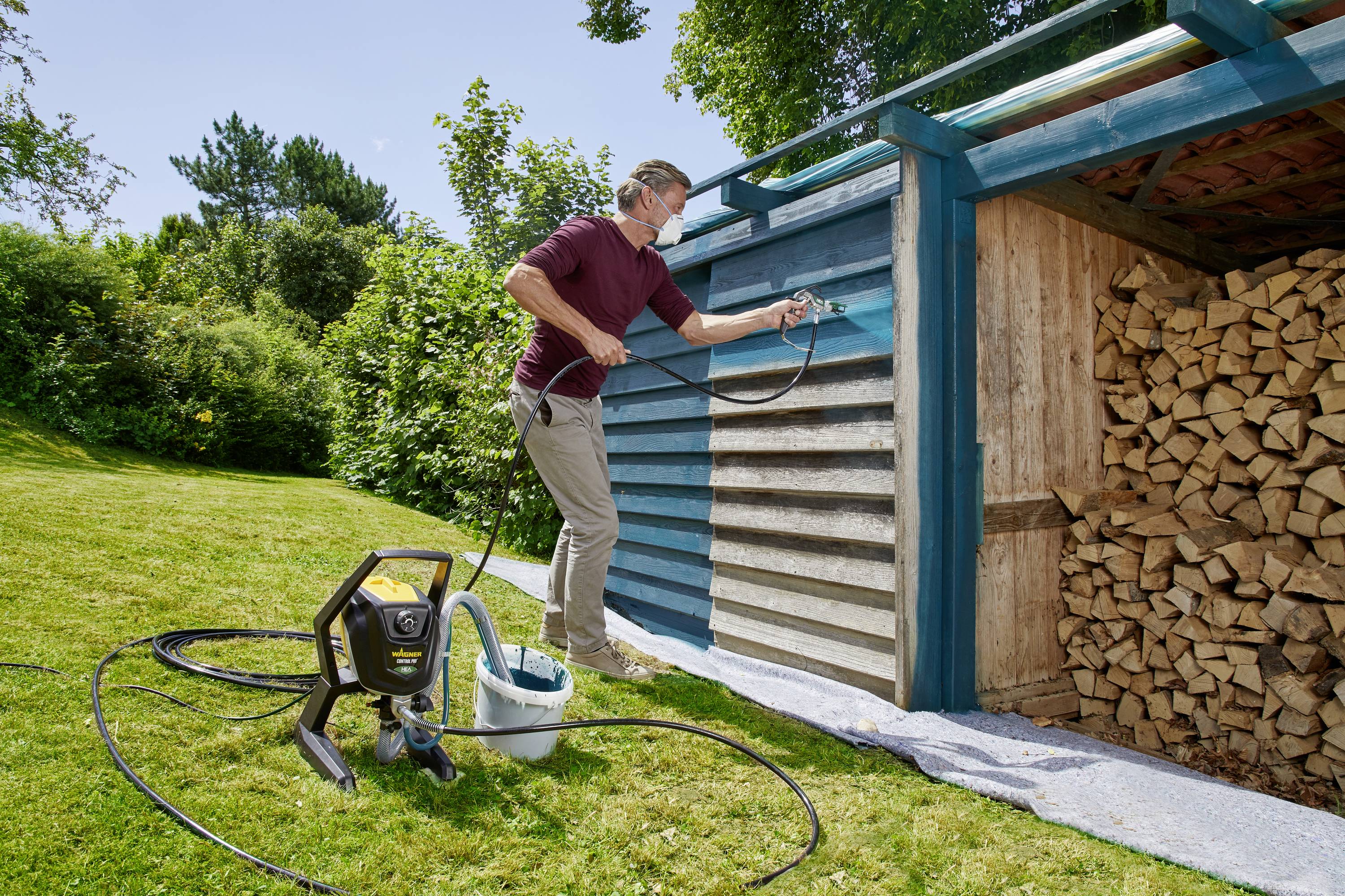 Eine Person lackiert ein Holzlager im Garten mit einem Farbsprüher. Links steht das Gerät, rechts ist das Holz gestapelt.