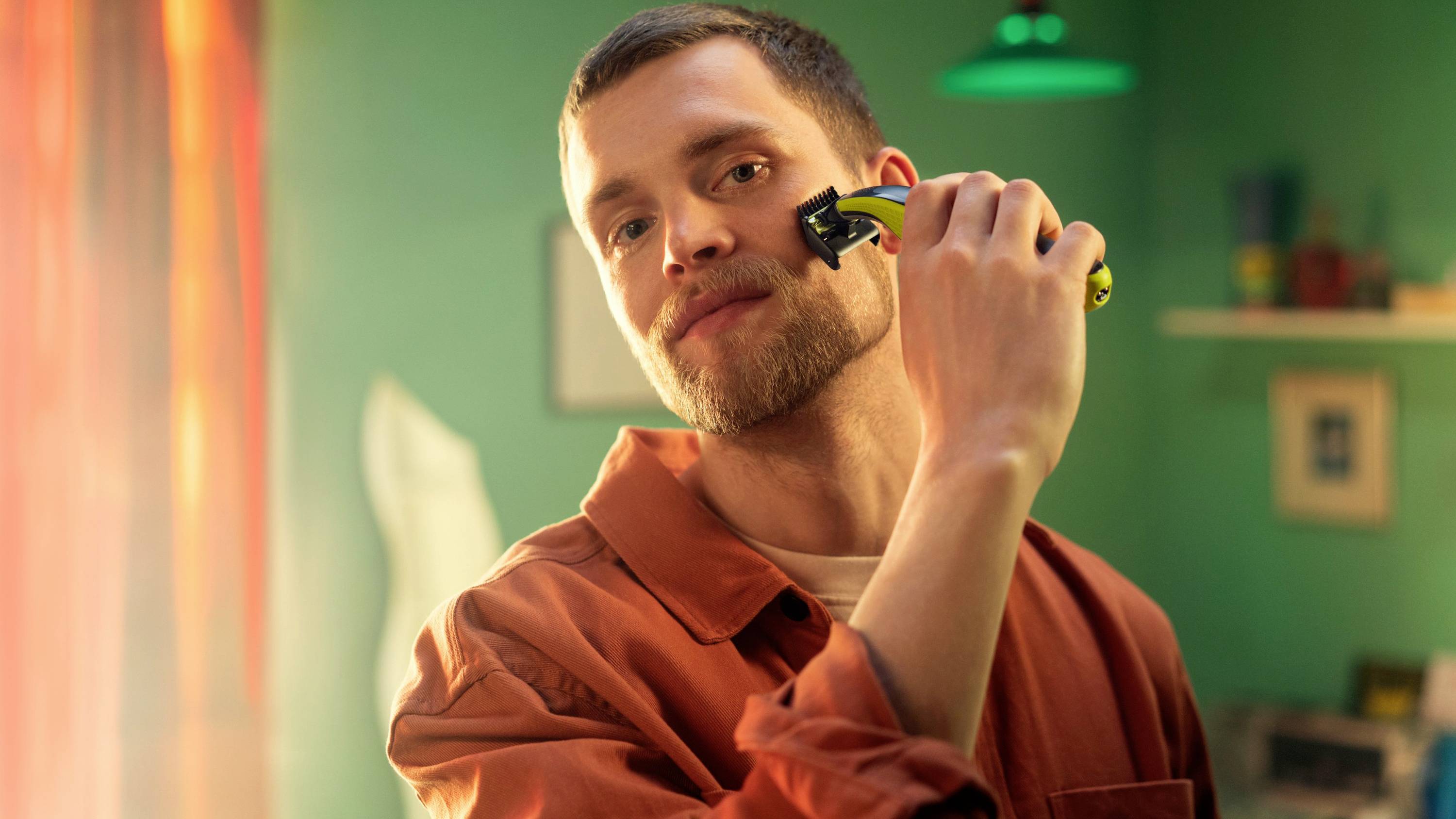 Ein Mann rasiert sich im Gesicht mit einem elektrischen Rasierer. Er befindet sich in einem Zimmer mit grünen Wänden und orangen Vorhängen.