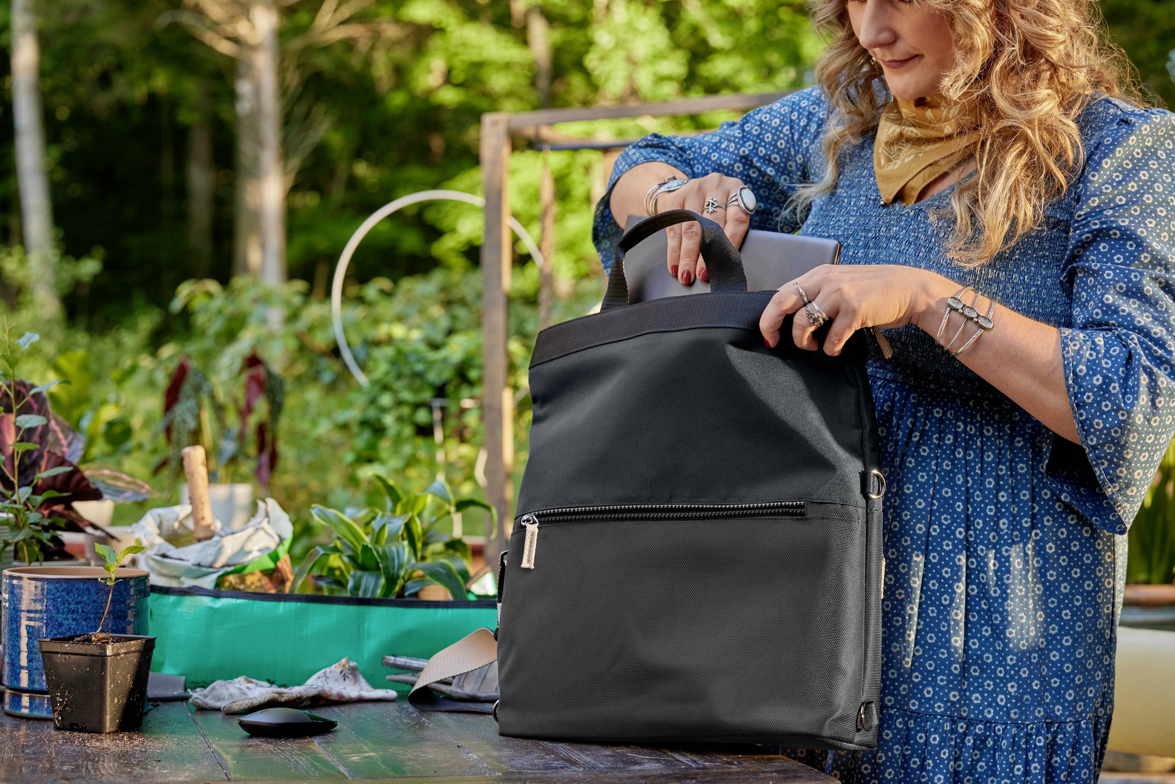 Frau in blauem Kleid packt schwarzen Rucksack an einem Holztisch im Garten. Auf dem Tisch stehen Pflanzen und Gartenwerkzeuge.