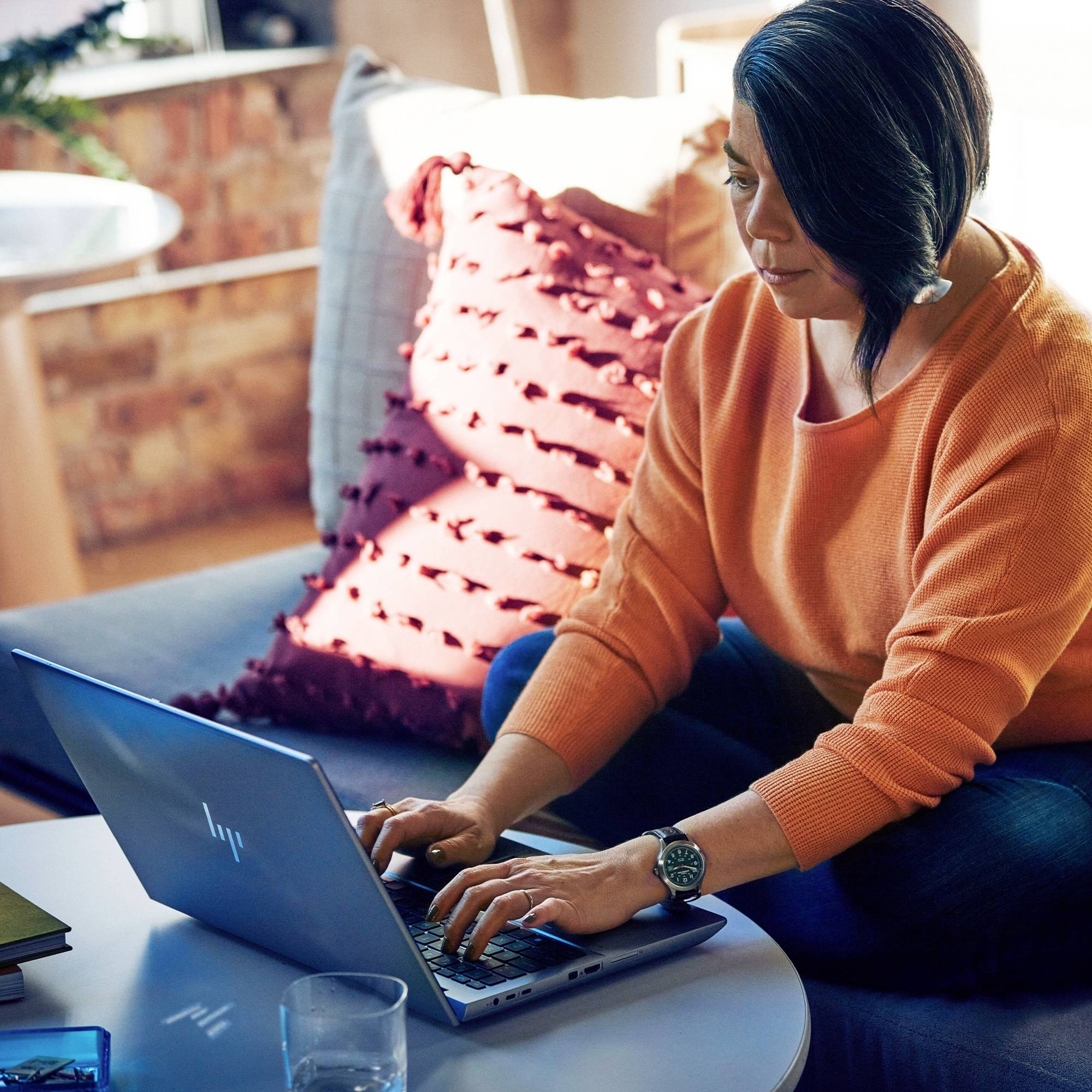 Eine Person sitzt in einem Wohnzimmer auf einem Sofa und tippt auf einem Laptop. Im Hintergrund sind Kissen und ein Fenster sichtbar.