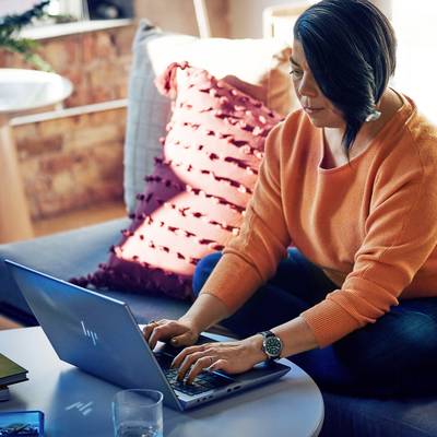 Eine Person sitzt auf einem Sofa und arbeitet an einem Laptop. Im Hintergrund sind Kissen und ein Fenster zu sehen.