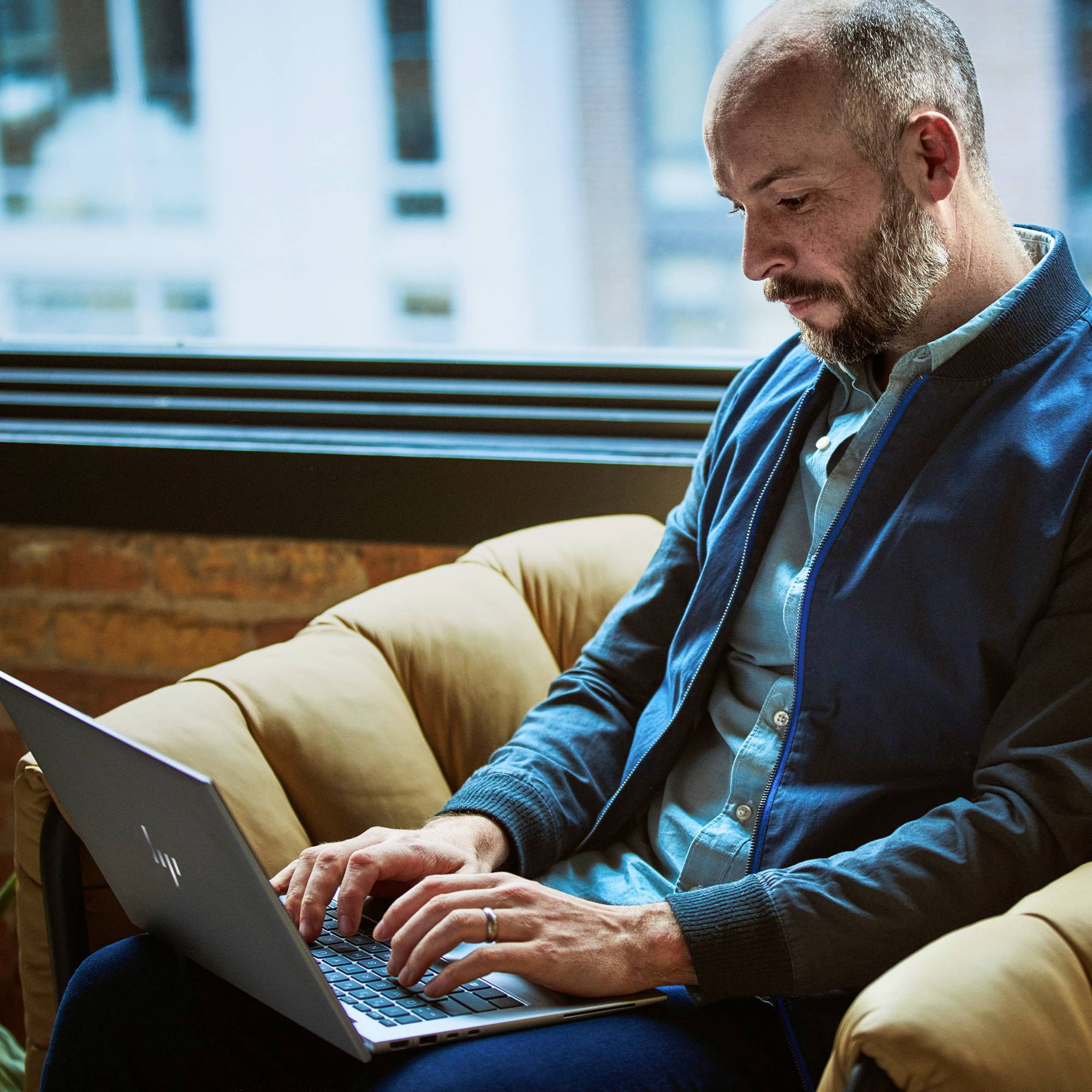 Mann sitzt auf einem Sofa, arbeitet an einem Laptop. Die Umgebung wirkt entspannt mit natürlichem Licht durch ein Fenster im Hintergrund.