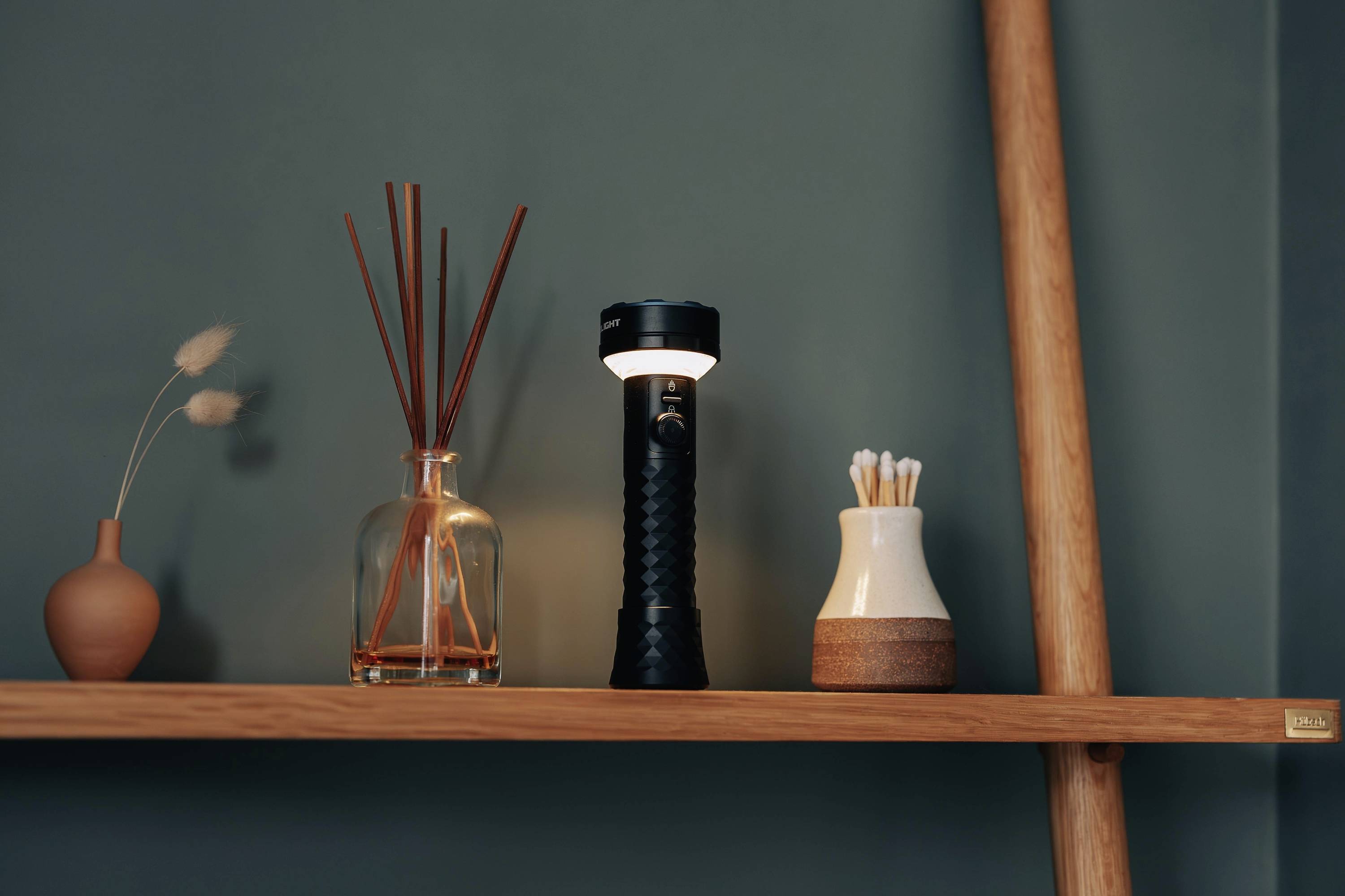Auf einem Holzregal stehen Dekorationsartikel: eine Vase, ein Diffuser, eine Taschenlampe und ein Behälter mit Wattestäbchen.