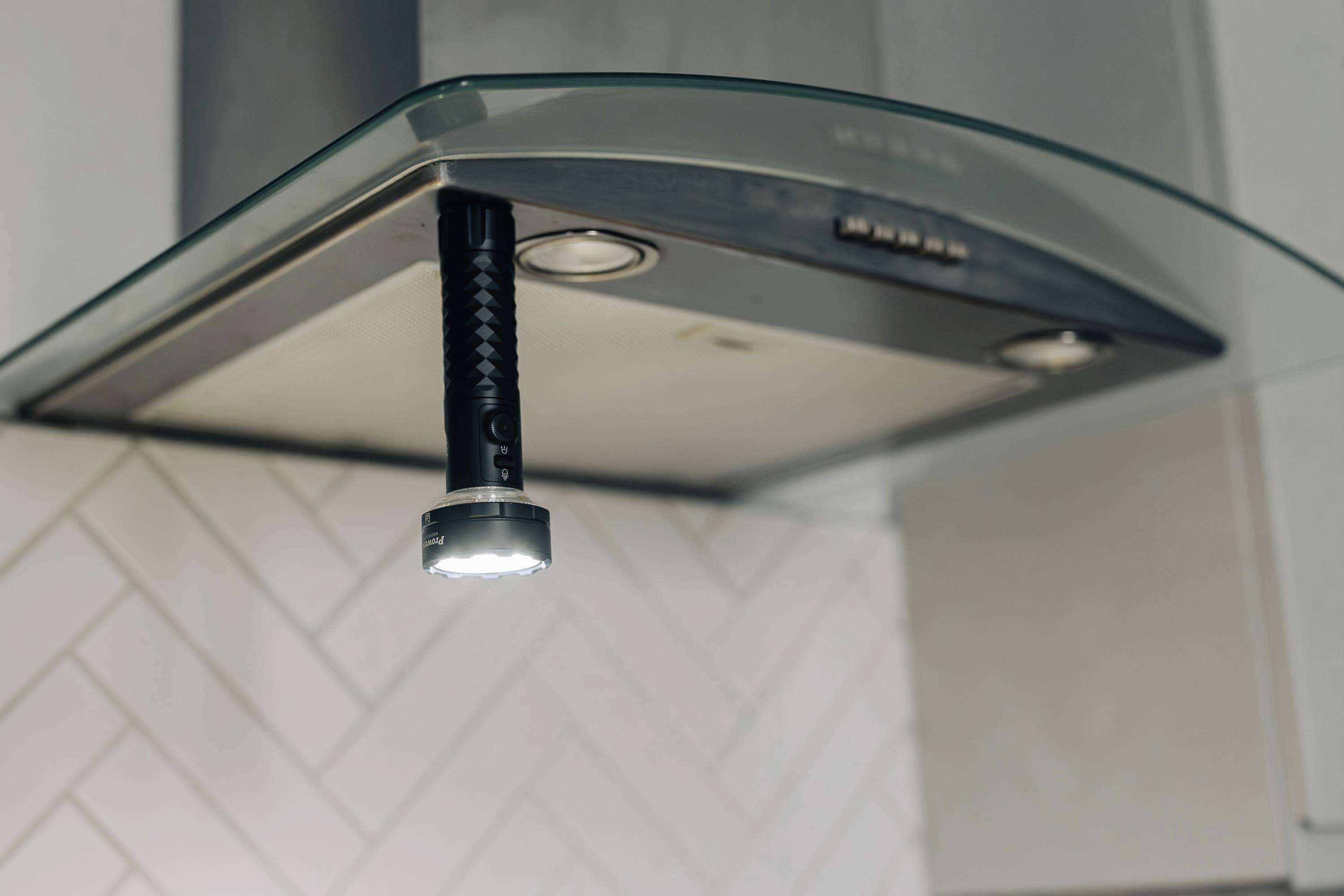 Eine schwarze Taschenlampe hängt magnetisch an einer Dunstabzugshaube in einer Küche. Die Beleuchtung ist eingeschaltet.