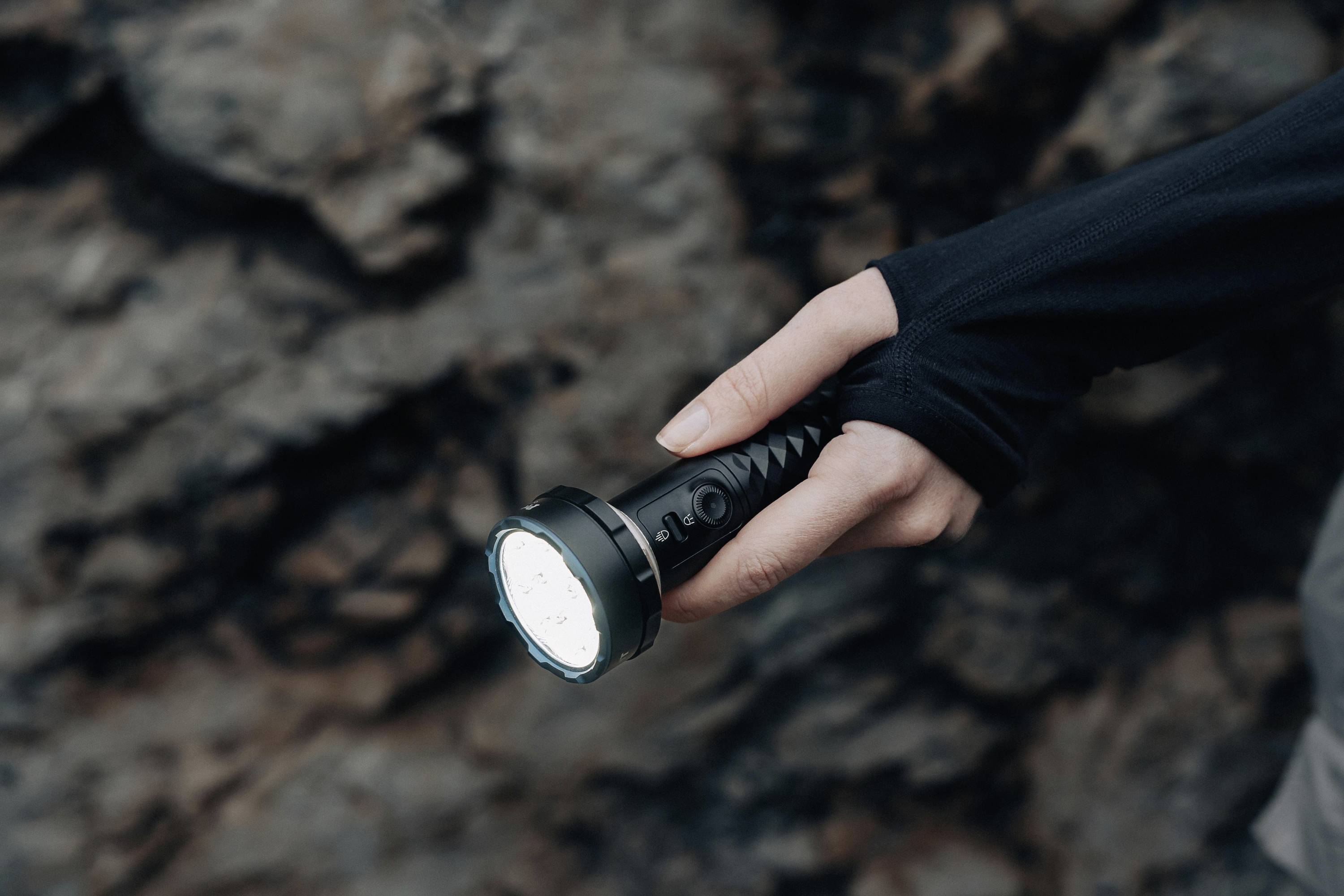 Eine Hand hält eine leuchtende Taschenlampe vor einem dunklen, felsigen Hintergrund, möglicherweise in einer Höhle oder bei Nacht.