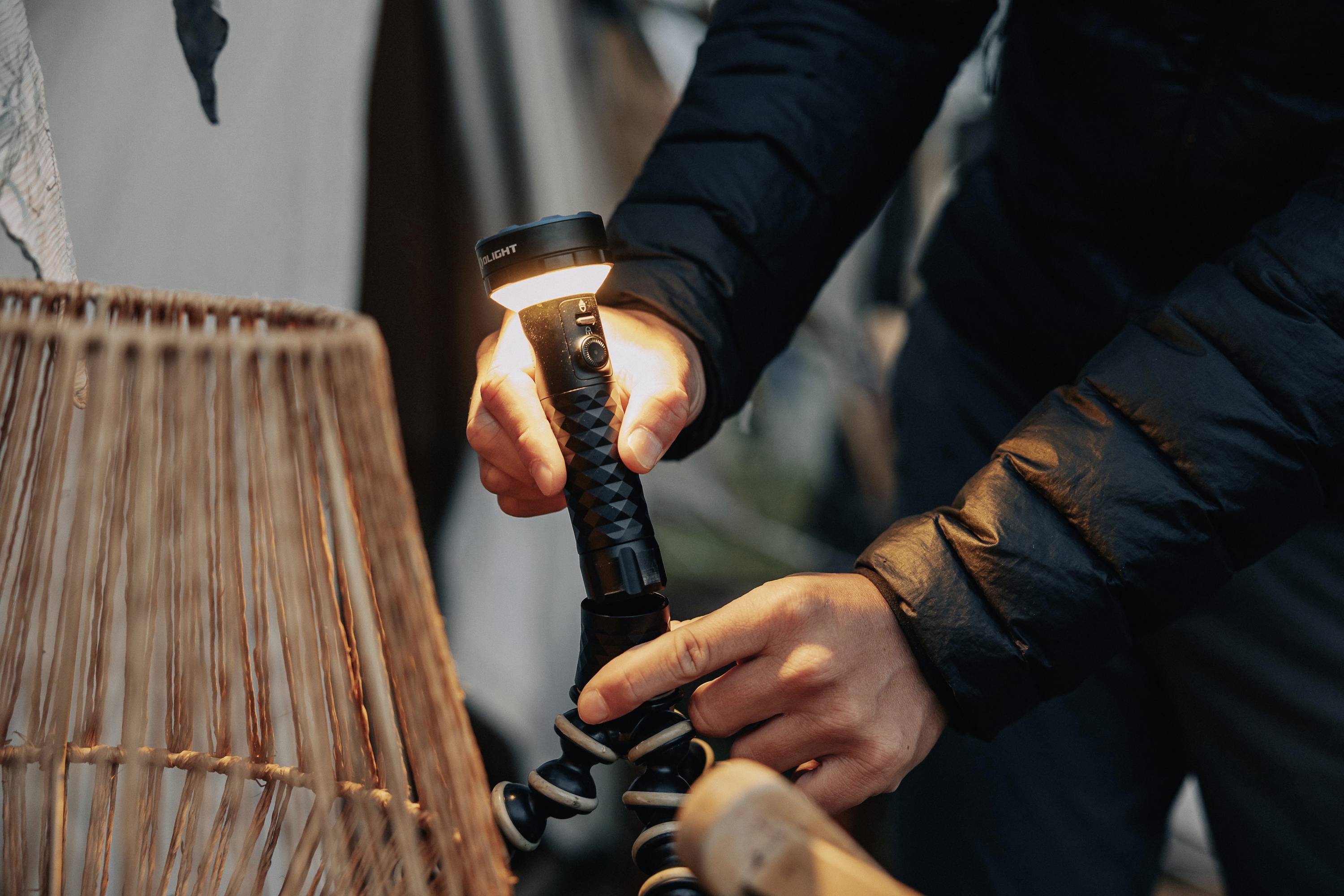 Eine Person in einer dunklen Jacke hält eine leuchtende Taschenlampe und befestigt sie auf einem flexiblen Stativ neben einem geflochtenen Lampenschirm.