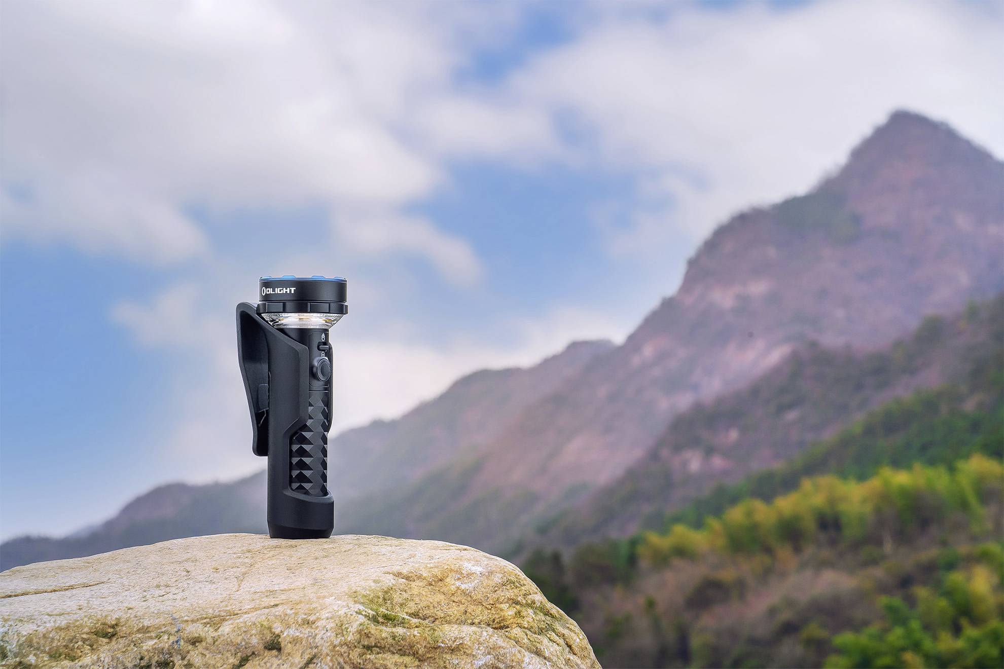 Eine Taschenlampe steht auf einem Felsen vor einer bergigen Landschaft mit bewölktem Himmel.
