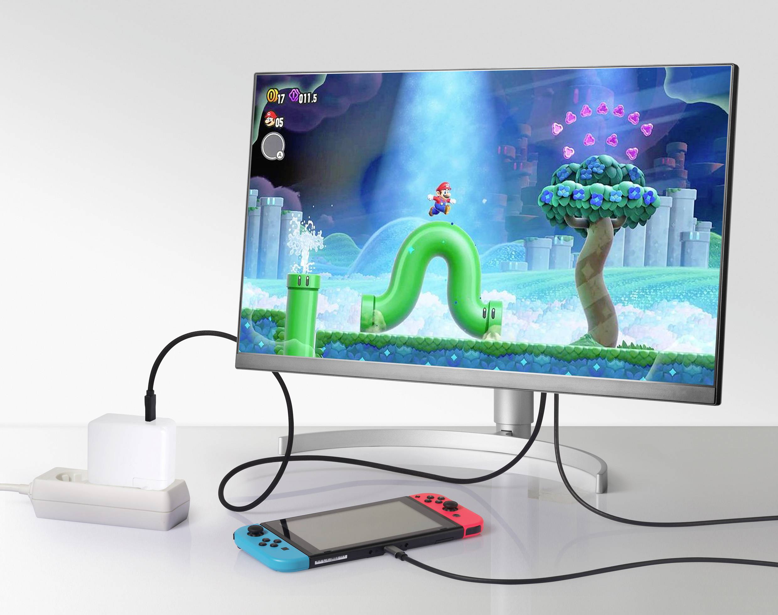 Ein Monitor zeigt ein Videospiel mit einer Spielfigur, die auf einer grünen Röhre steht, umgeben von Wasser und Bäumen. Darunter eine angeschlossene Konsole.