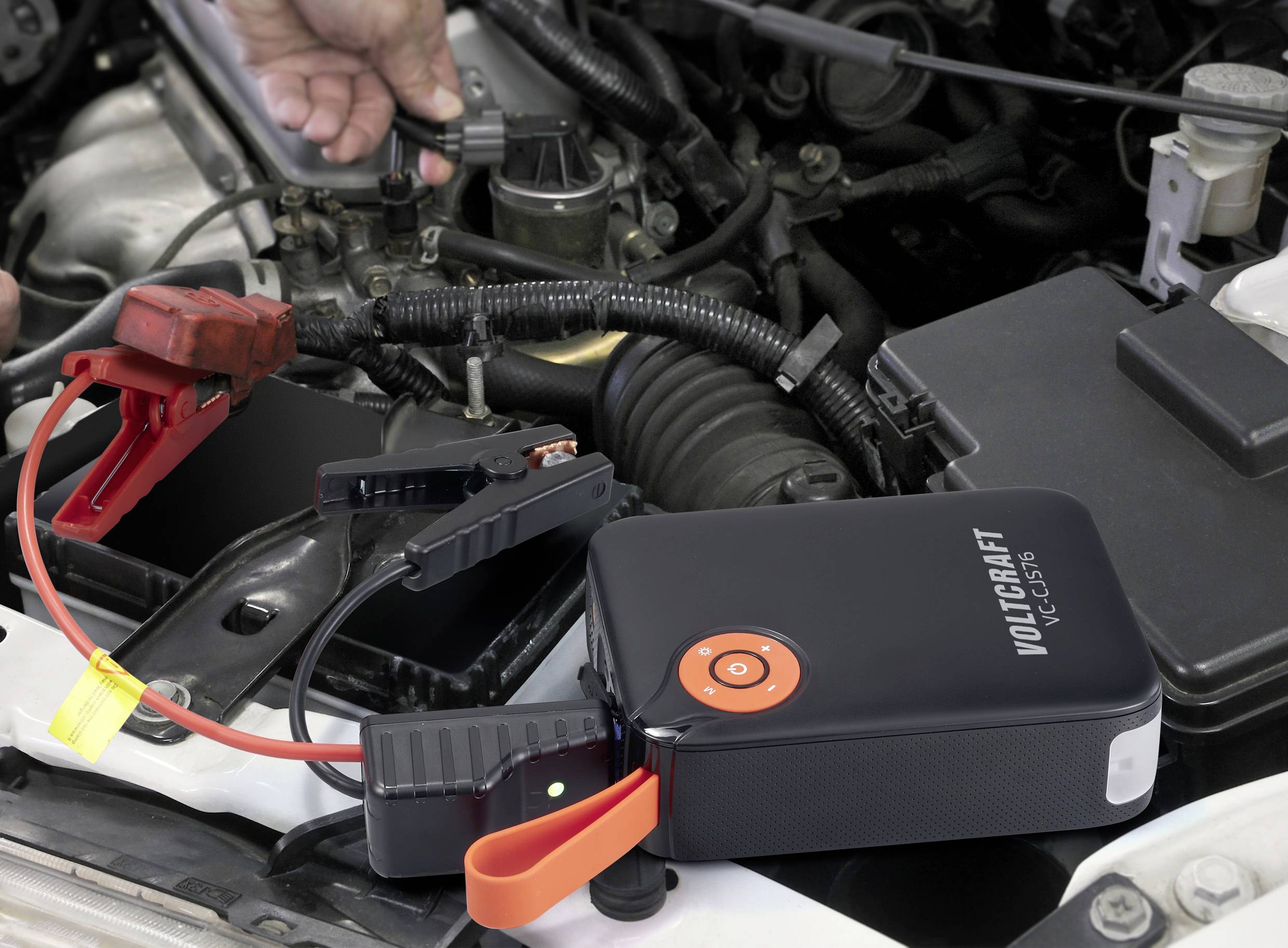Auto-Batterieladegerät wird an eine Fahrzeugbatterie angeschlossen. Hände halten Kabel, die Motorhaube ist offen.
