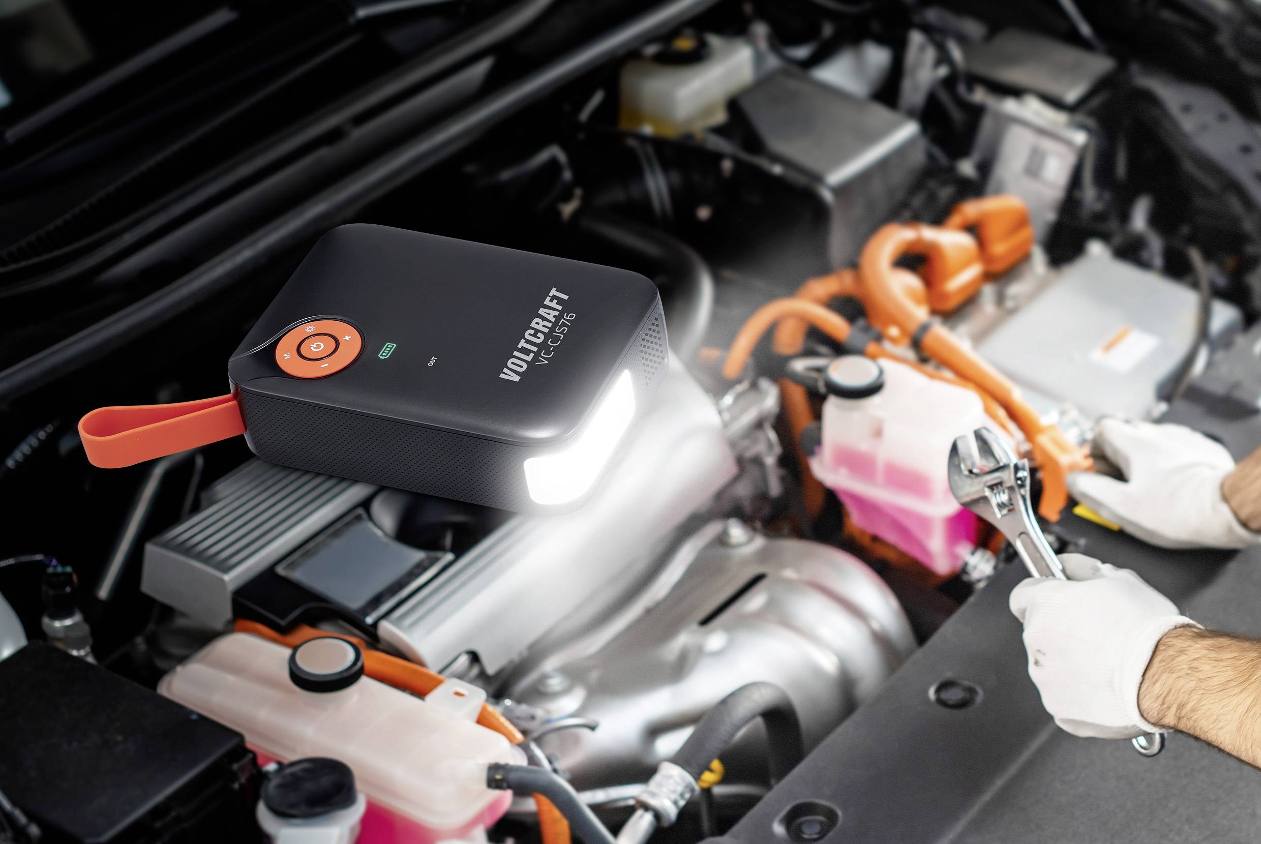 Eine Person repariert einen Motor. Ein tragbares Starthilfe-Gerät leuchtet den Motorraum aus.