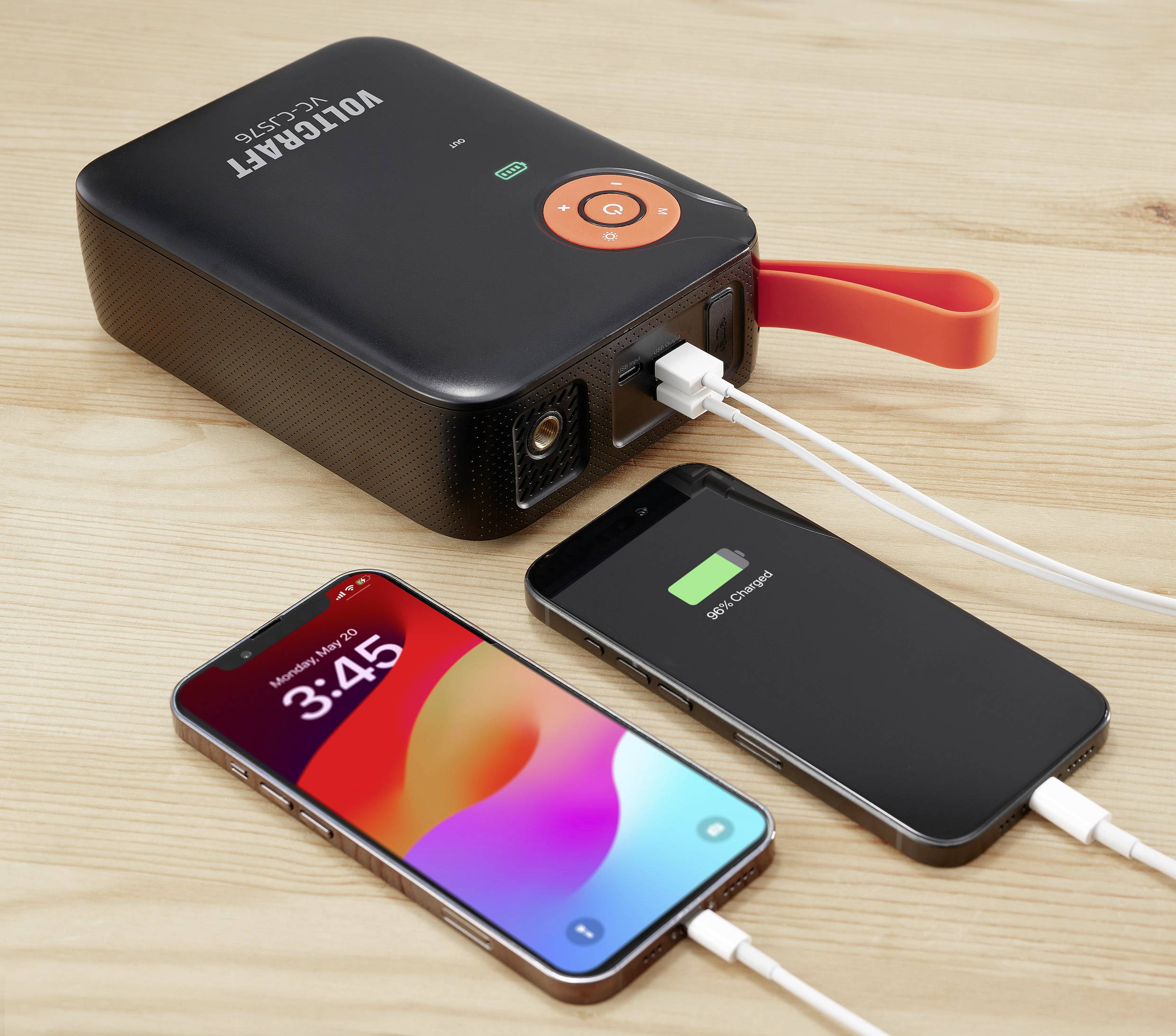 Ein tragbares Ladegerät lädt zwei Handys auf einem Holztisch. Ein Handy zeigt eine Uhrzeit an, das andere zeigt einen vollen Akku.