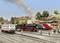 'Eisenbahnmodellanlage mit mehreren Zügen: Dampflok mit Rauch und moderner Zug im Hintergrund. Menschen auf dem Bahnsteig.'