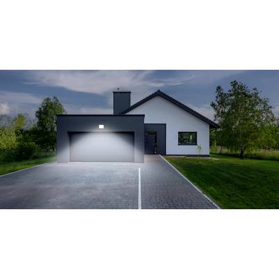 Ein modernes Einfamilienhaus mit weißer Fassade, grauem Dach und beleuchteter Einfahrt, umgeben von Bäumen und Rasen im Abendlicht.