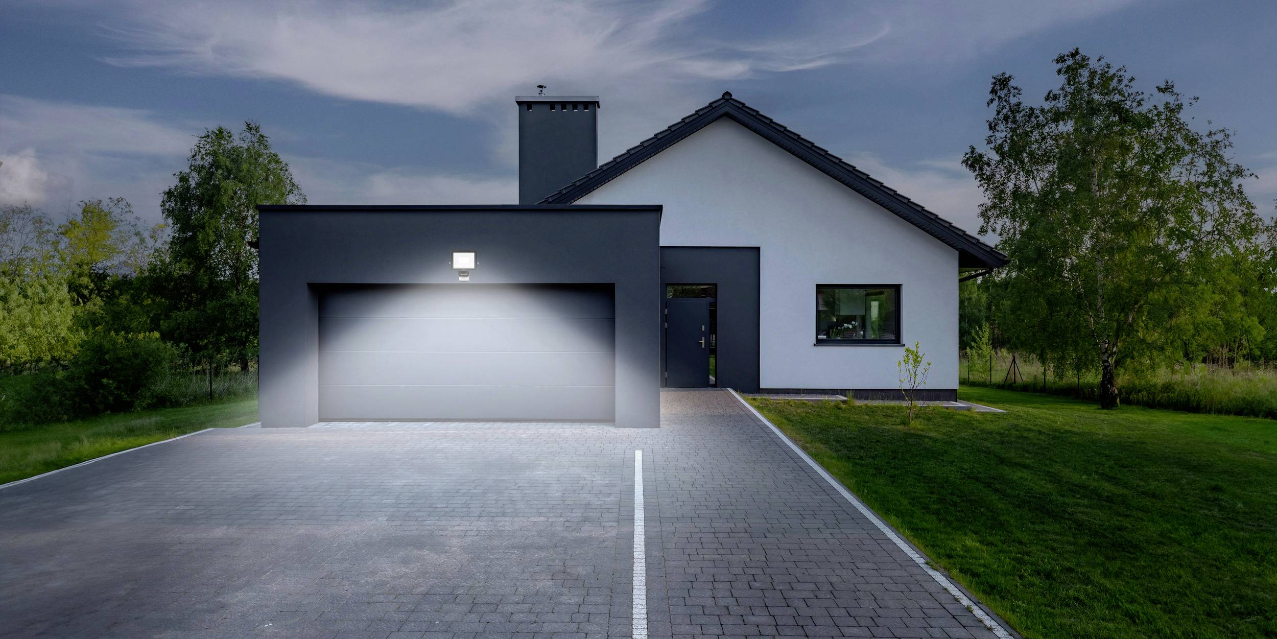 Ein modernes Einfamilienhaus mit beleuchteter Einfahrt und gepflegtem Garten, umgeben von Bäumen bei Dämmerung.