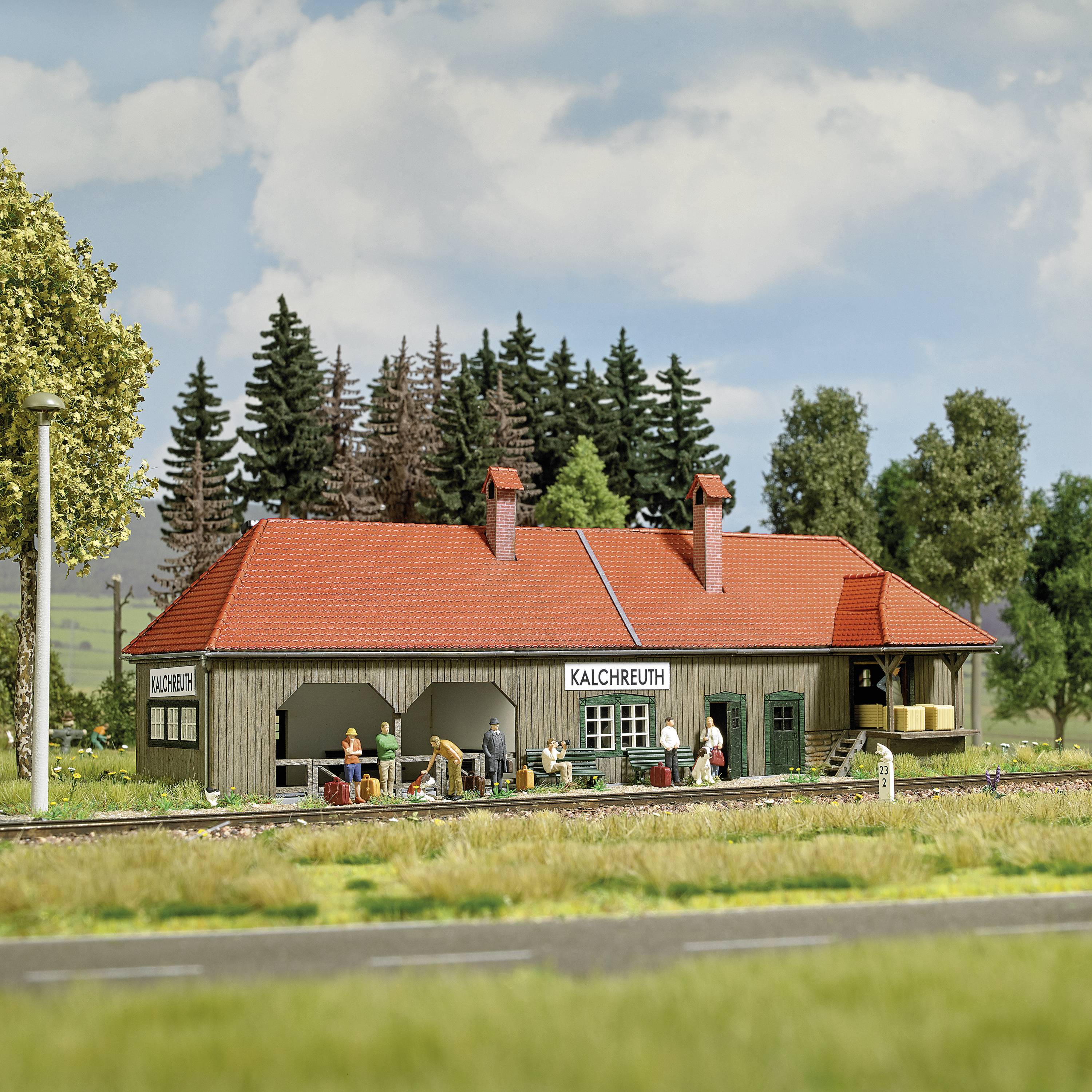Kleiner Bahnhof mit rotem Dach und Schild 'Kalchreuth', umgeben von Bäumen. Modelleisenbahn mit Figuren und Güterwagen im Vordergrund.