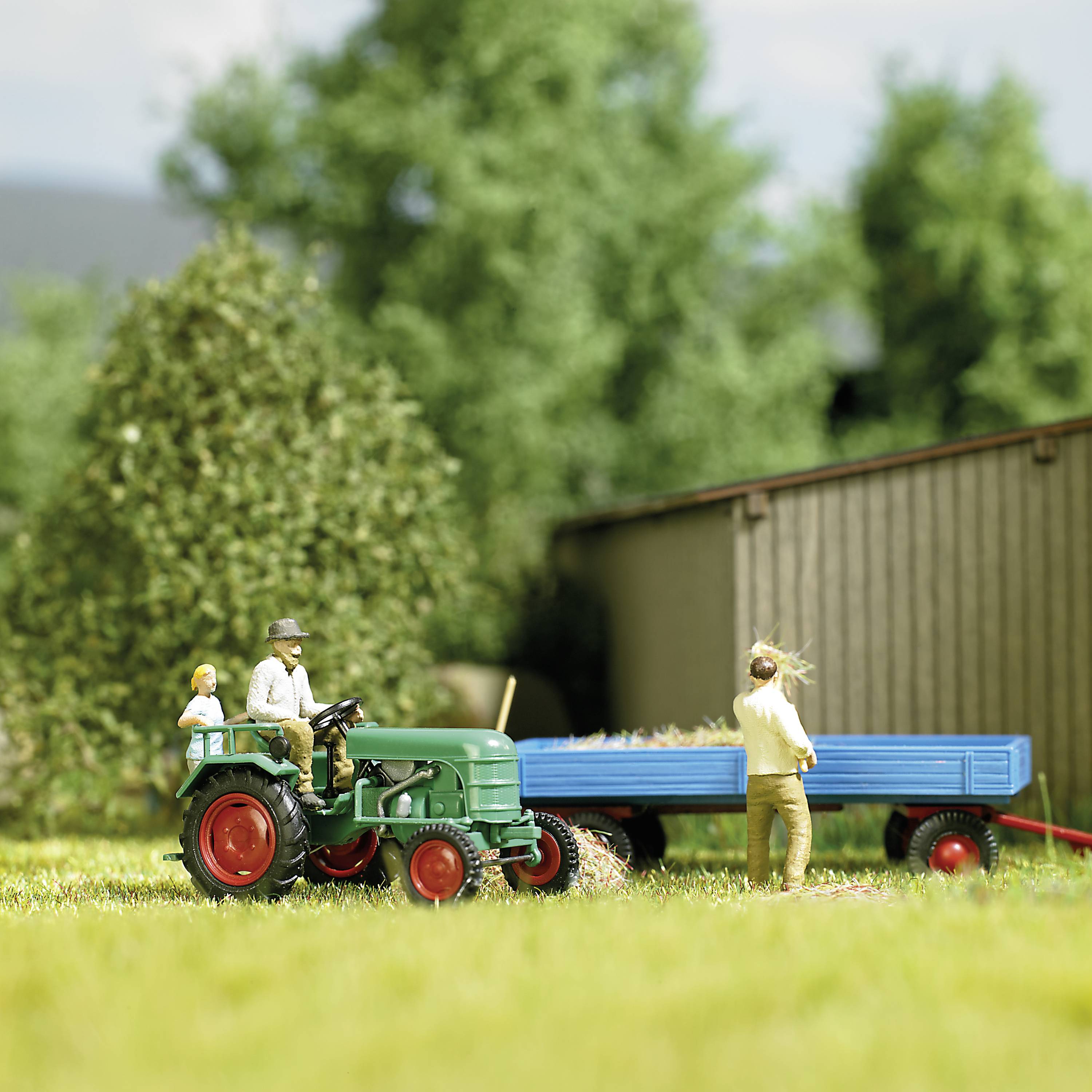 Ein Modelltraktor zieht einen Anhänger mit blauen Stangen über eine grüne Wiese. Im Hintergrund sind Bäume und ein Gebäude.