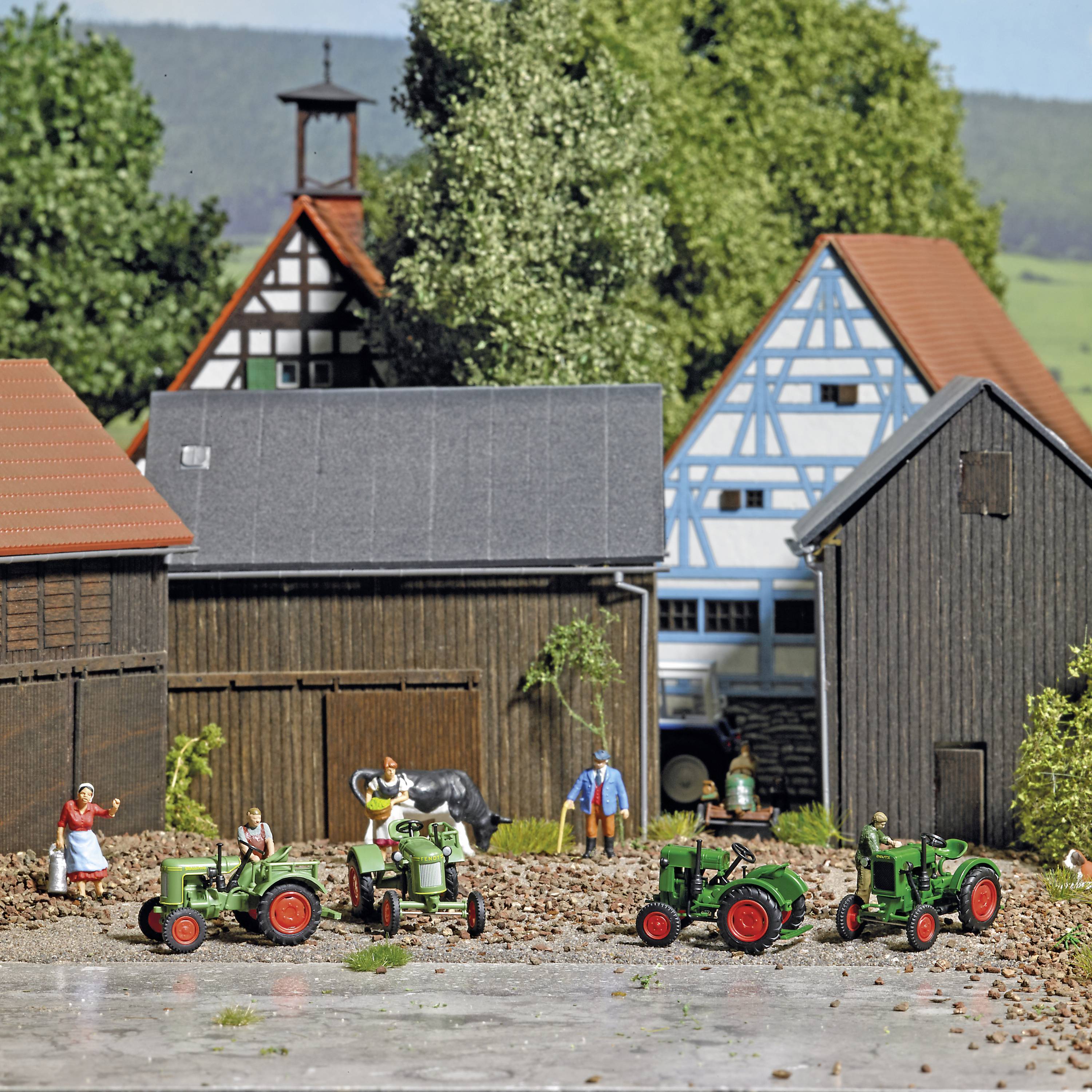 Drei grüne Traktoren stehen auf einem Bauernhof mit traditionellen Fachwerkhäusern im Hintergrund. Zwei Figuren interagieren neben den Traktoren.