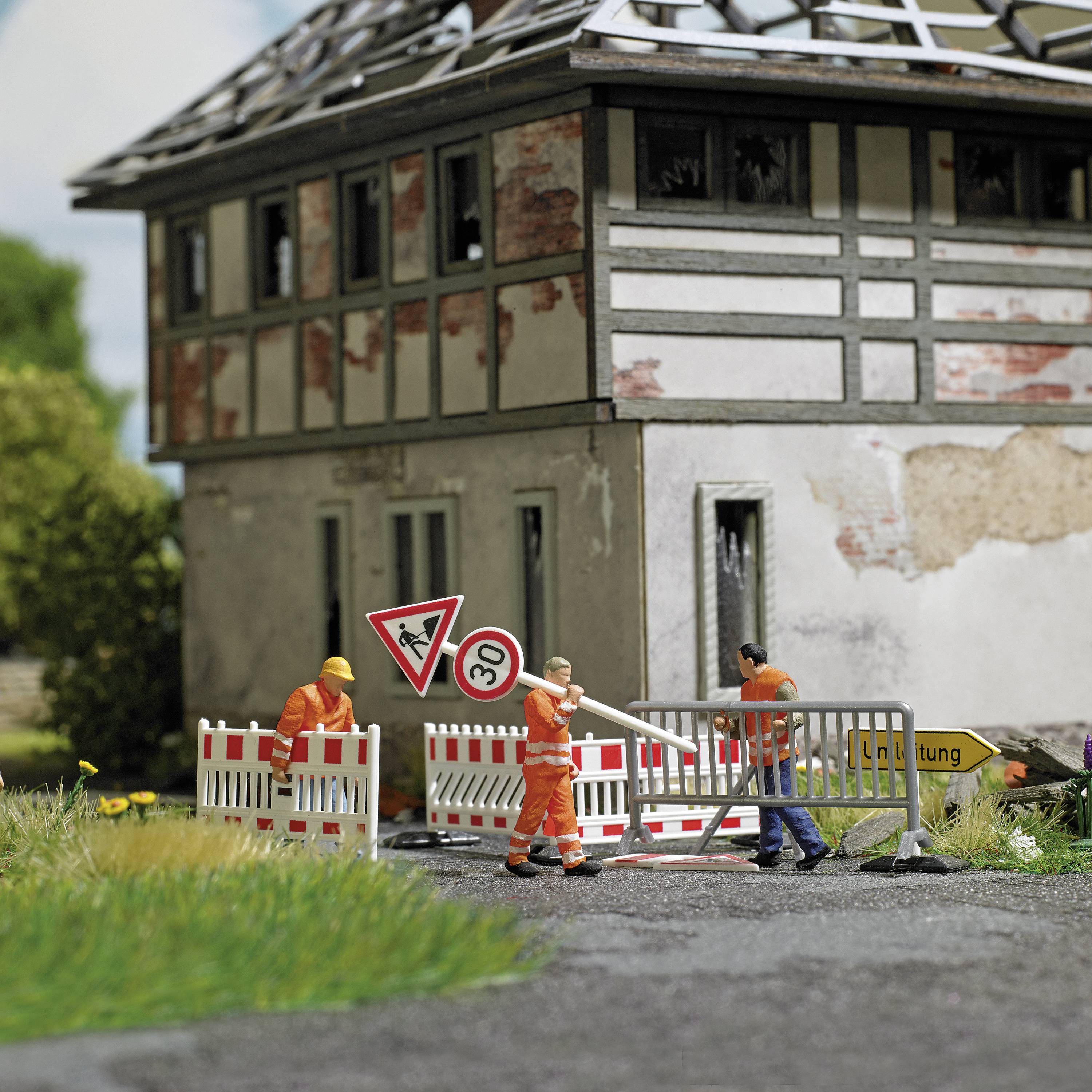 Arbeiter in orangefarbener Kleidung reparieren eine Straße vor einem alten Gebäude. Verkehrszeichen weisen auf Bauarbeiten hin.
