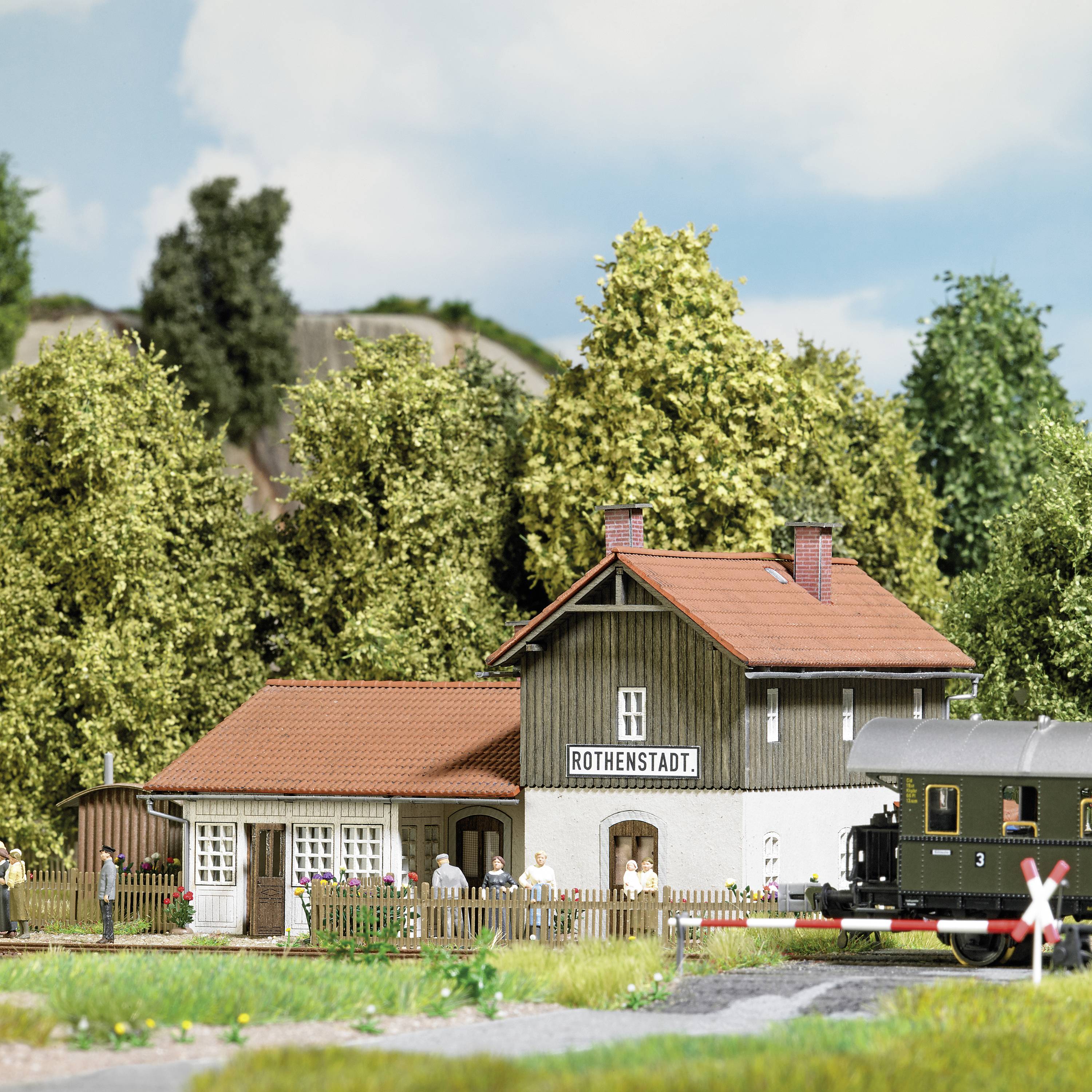 Ein Modellbahnhof mit der Beschriftung 'Rothenstadt', umgeben von grünen Bäumen und einer vorbeifahrenden Modellbahn.
