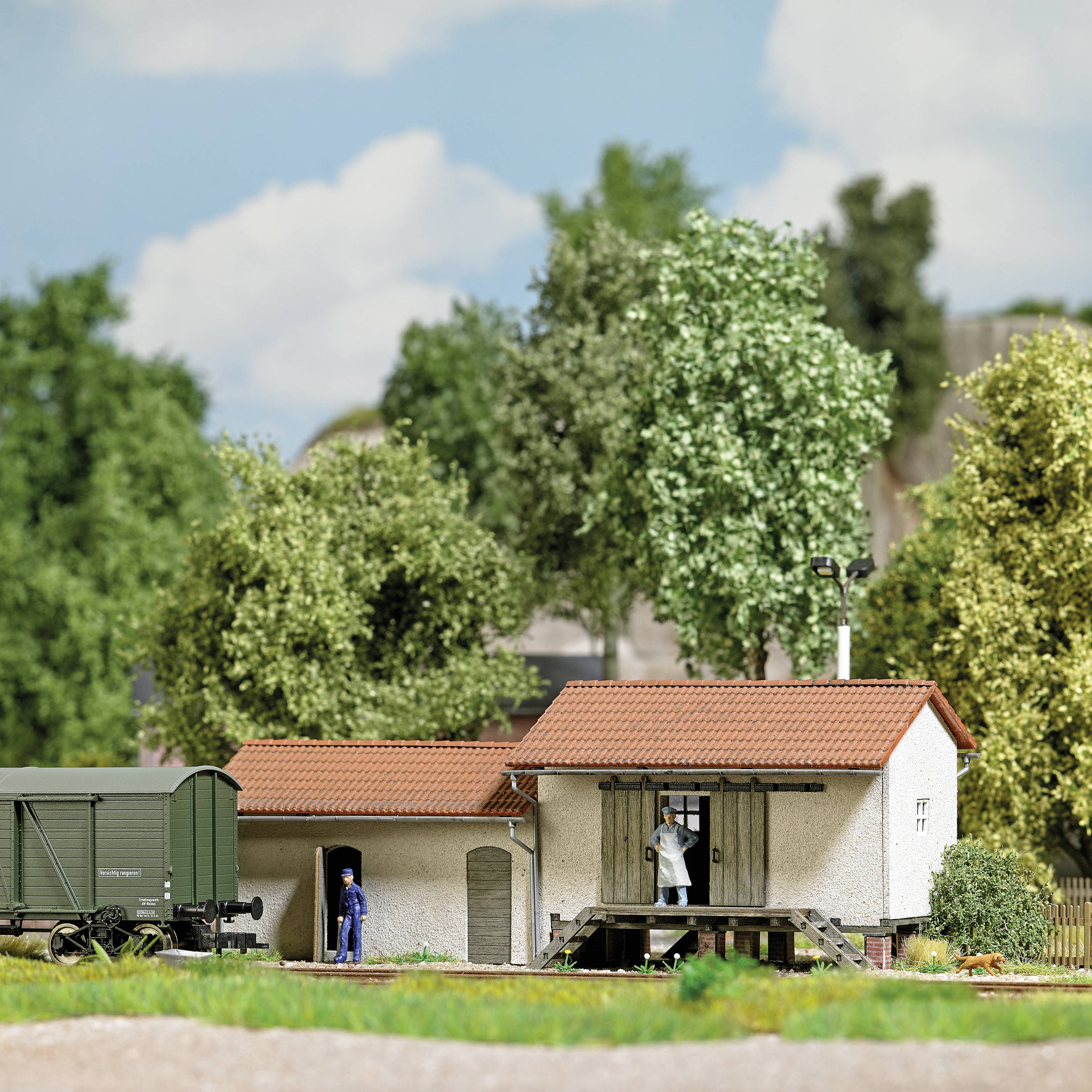 Kleiner Bahnhof im Grünen mit Schienen, Güterwagen und zwei Modellfiguren. Hintergrund zeigt Bäume und blauen Himmel.