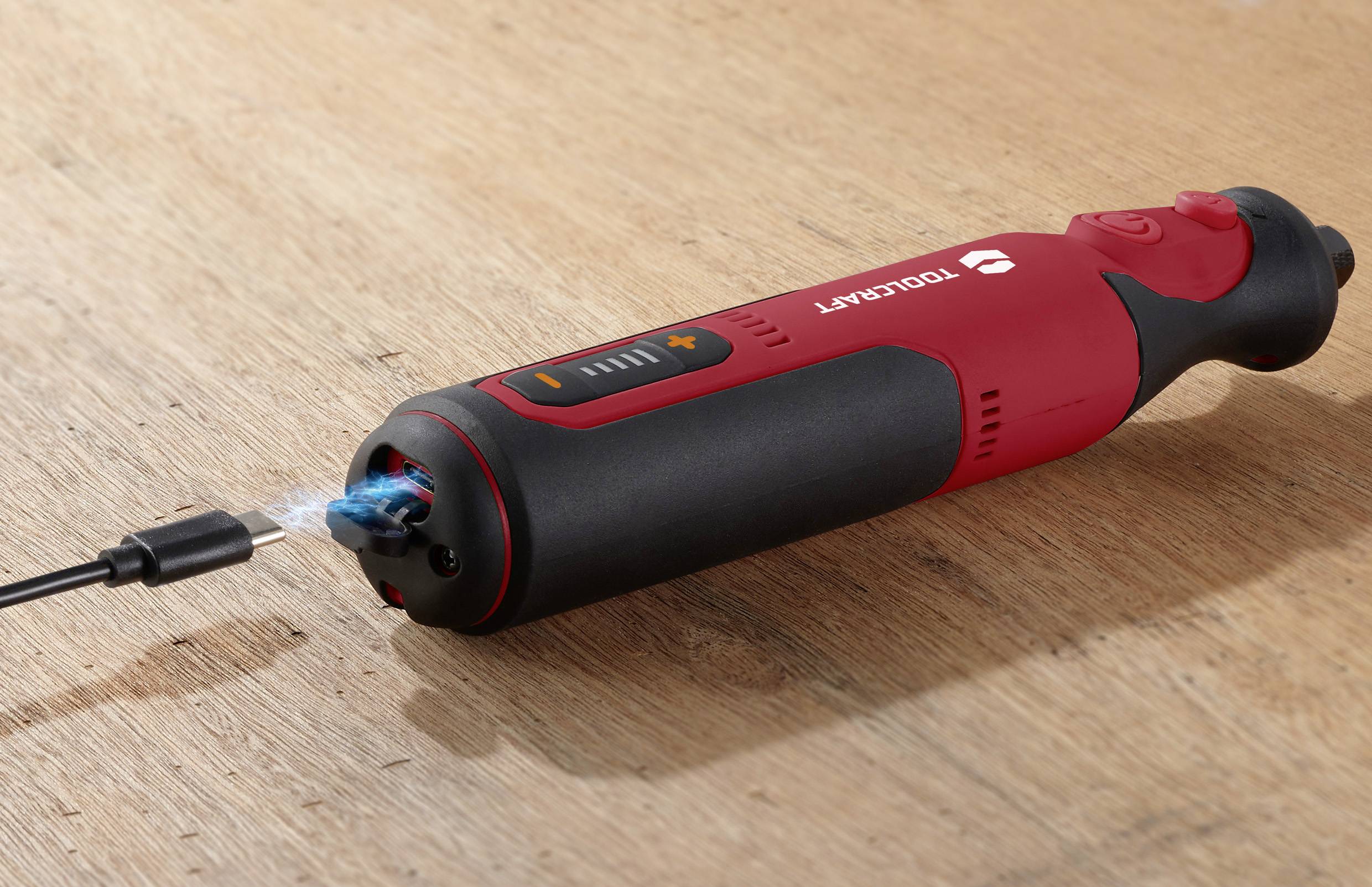 Ein roter, kabelloser Elektroschrauber liegt auf einem Holztisch. Ein USB-Kabel ist angeschlossen und lädt das Gerät auf.