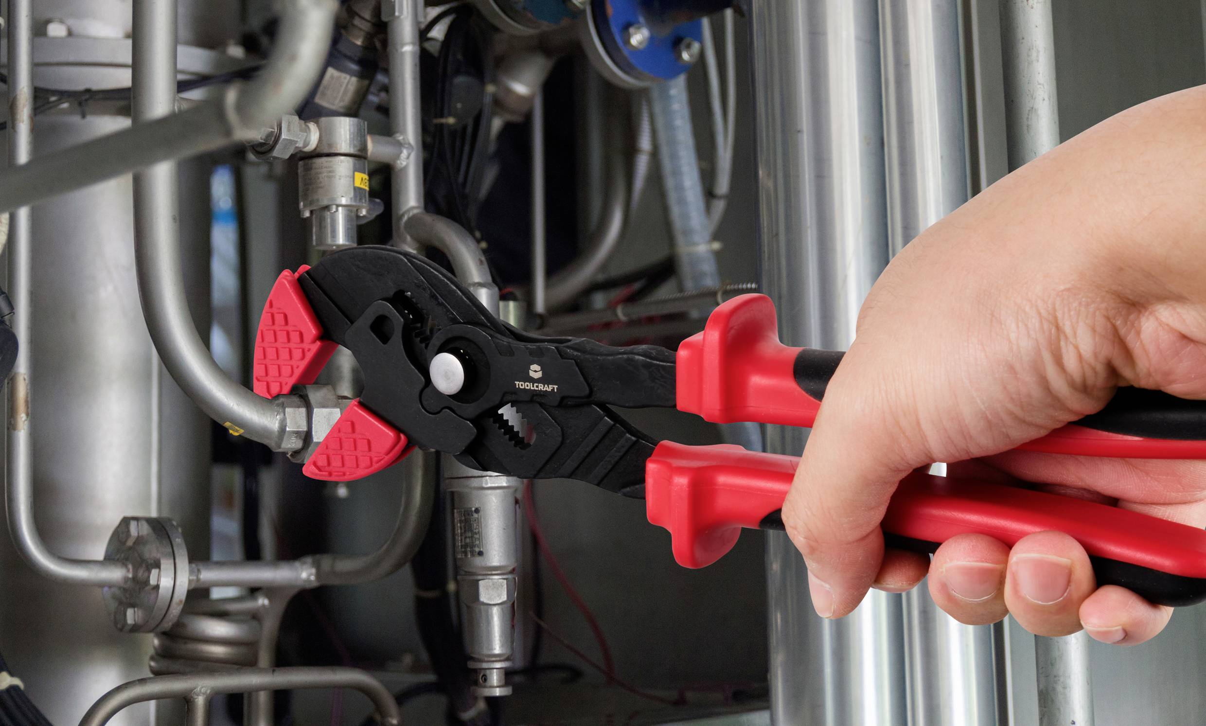 Eine Hand hält eine rote Rohrzange, die an einer Metallleitung arbeitet. Umgebende Rohrleitungen und Ventile sind zu sehen.