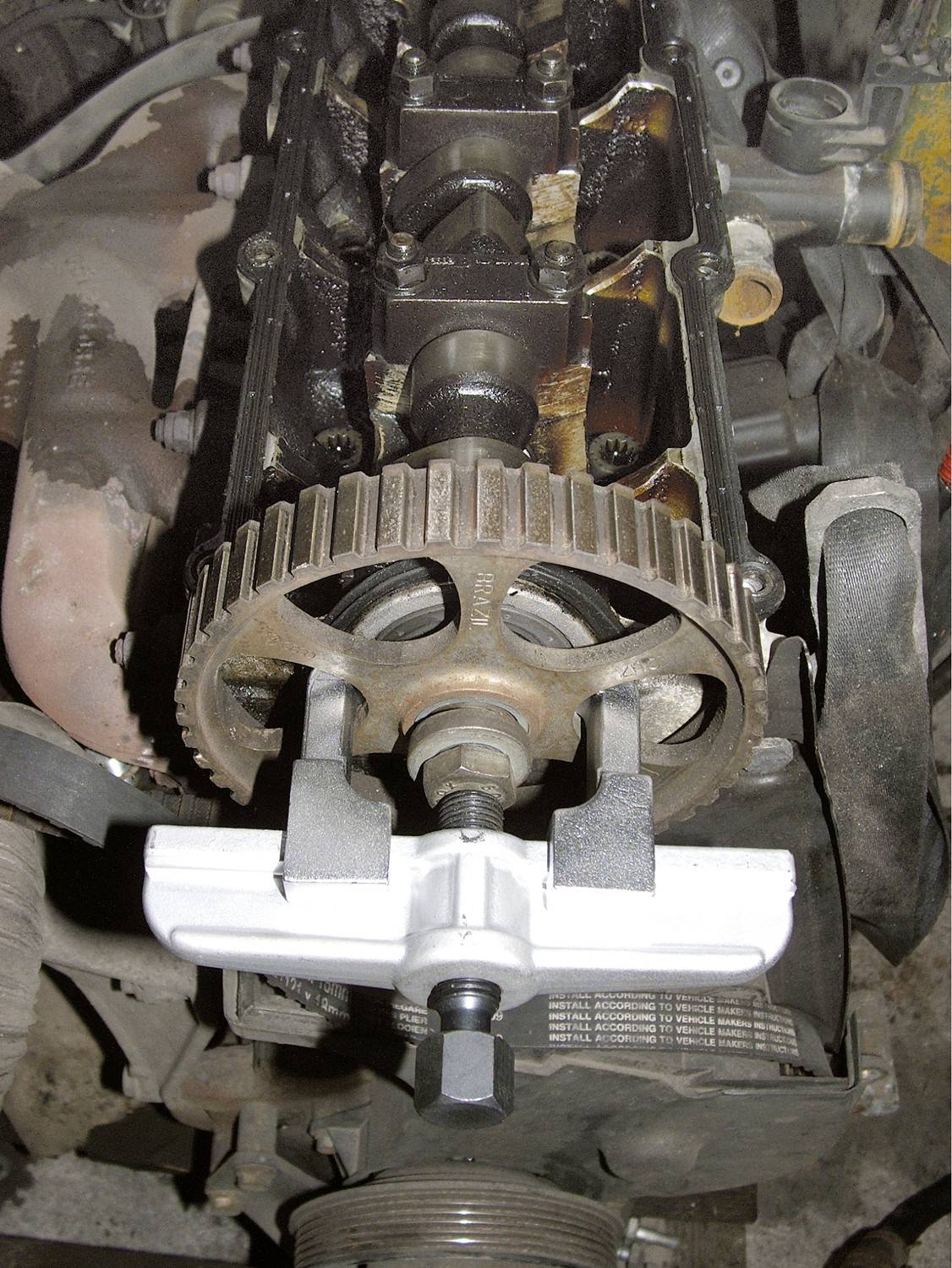 Offener Motor eines Autos mit Zahnriemenrad und Spannvorrichtung. Fokus liegt auf mechanischen Details der Motorstruktur.