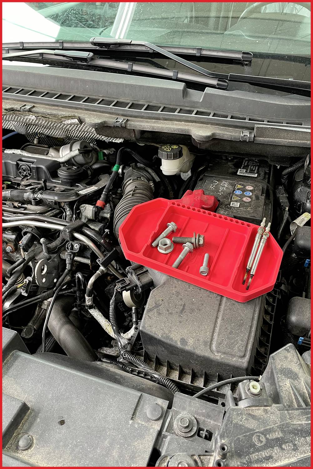 Ein offener Motorraum eines Autos mit einer roten Werkzeugablage auf der Batterie. Verschiedene Schrauben und Werkzeuge sind sichtbar.