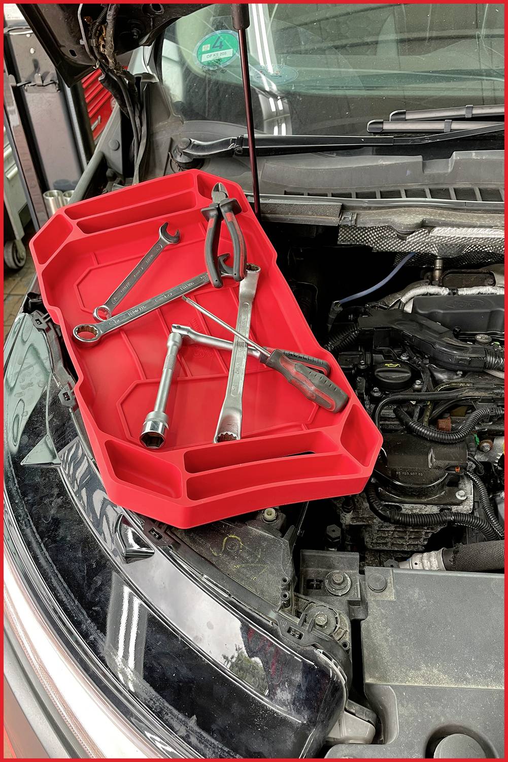 Roter Werkzeugkasten mit Schraubenschlüsseln und Zange auf einer geöffneten Motorhaube in einer Werkstatt.